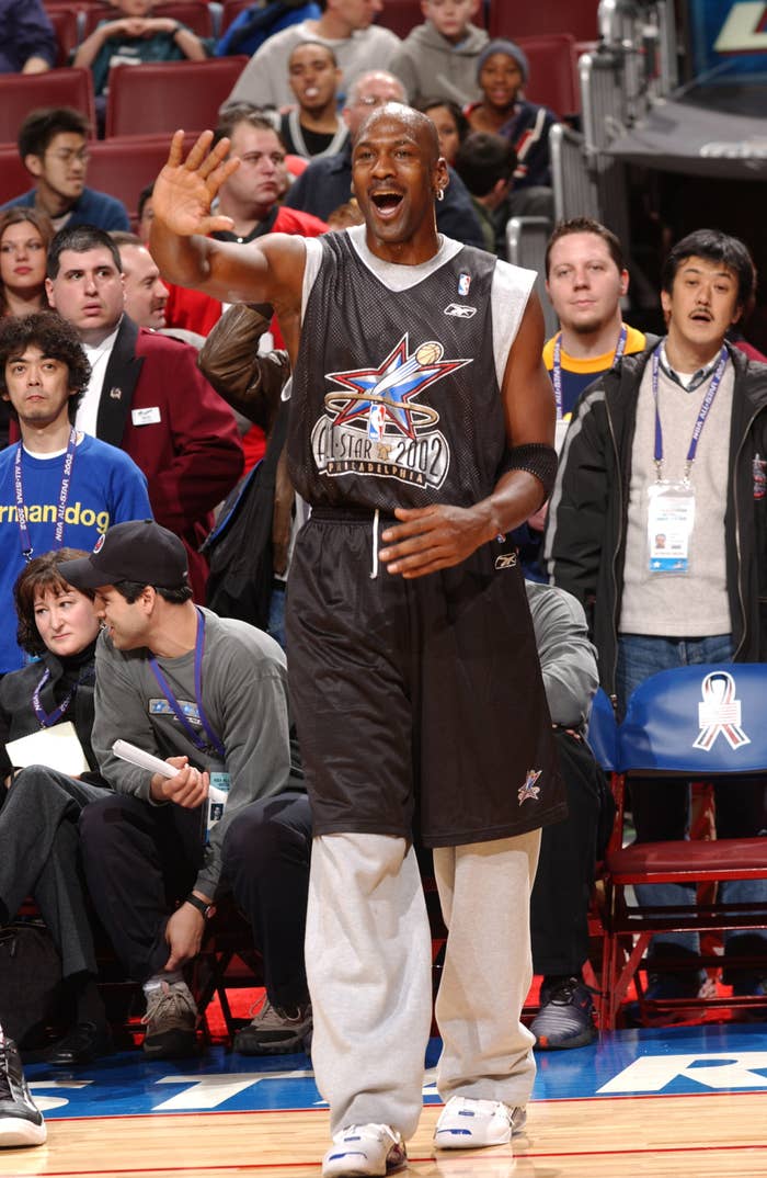 Basketball player wearing a 2002 All-Star jersey waves excitedly, standing on a basketball court surrounded by media and fans