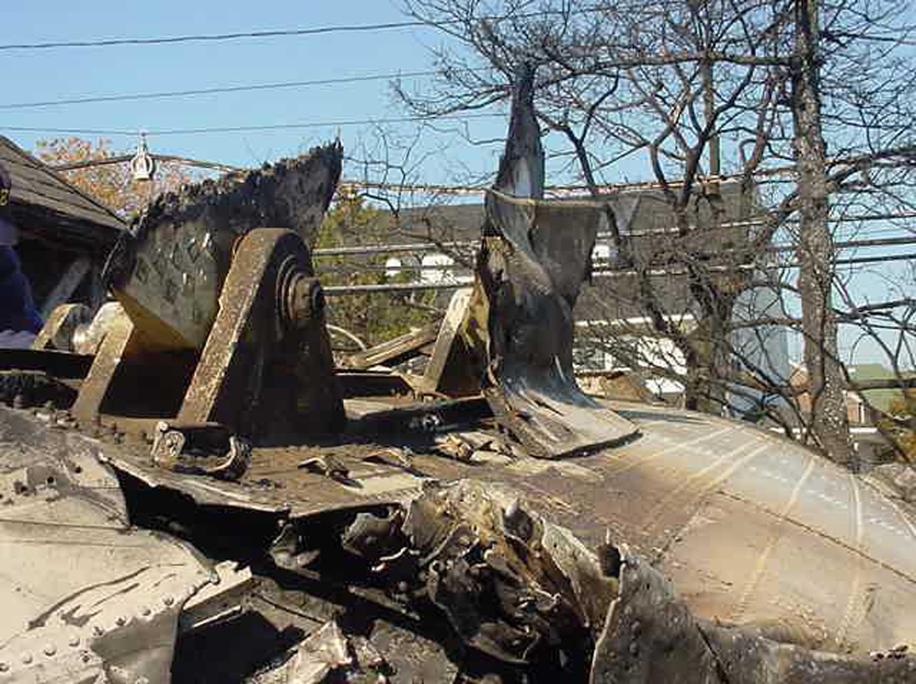 Wreckage of a destroyed Ferris wheel, partially burnt, in a wooded area with power lines visible in the background