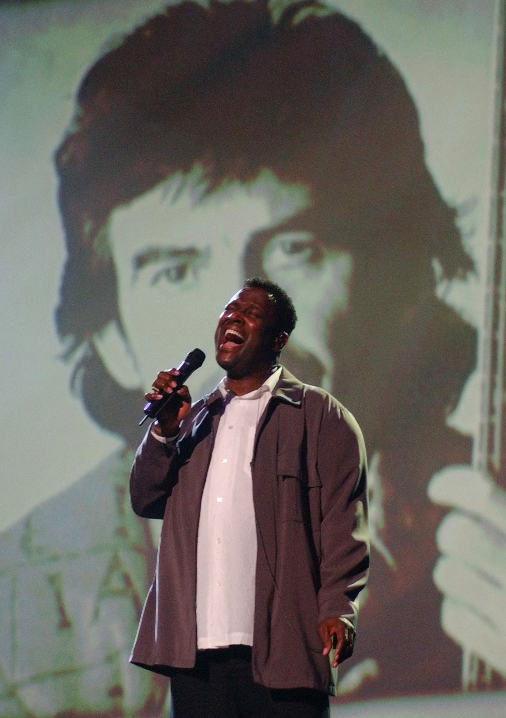 A person passionately sings on stage, holding a microphone, with a large projected image of another person behind them