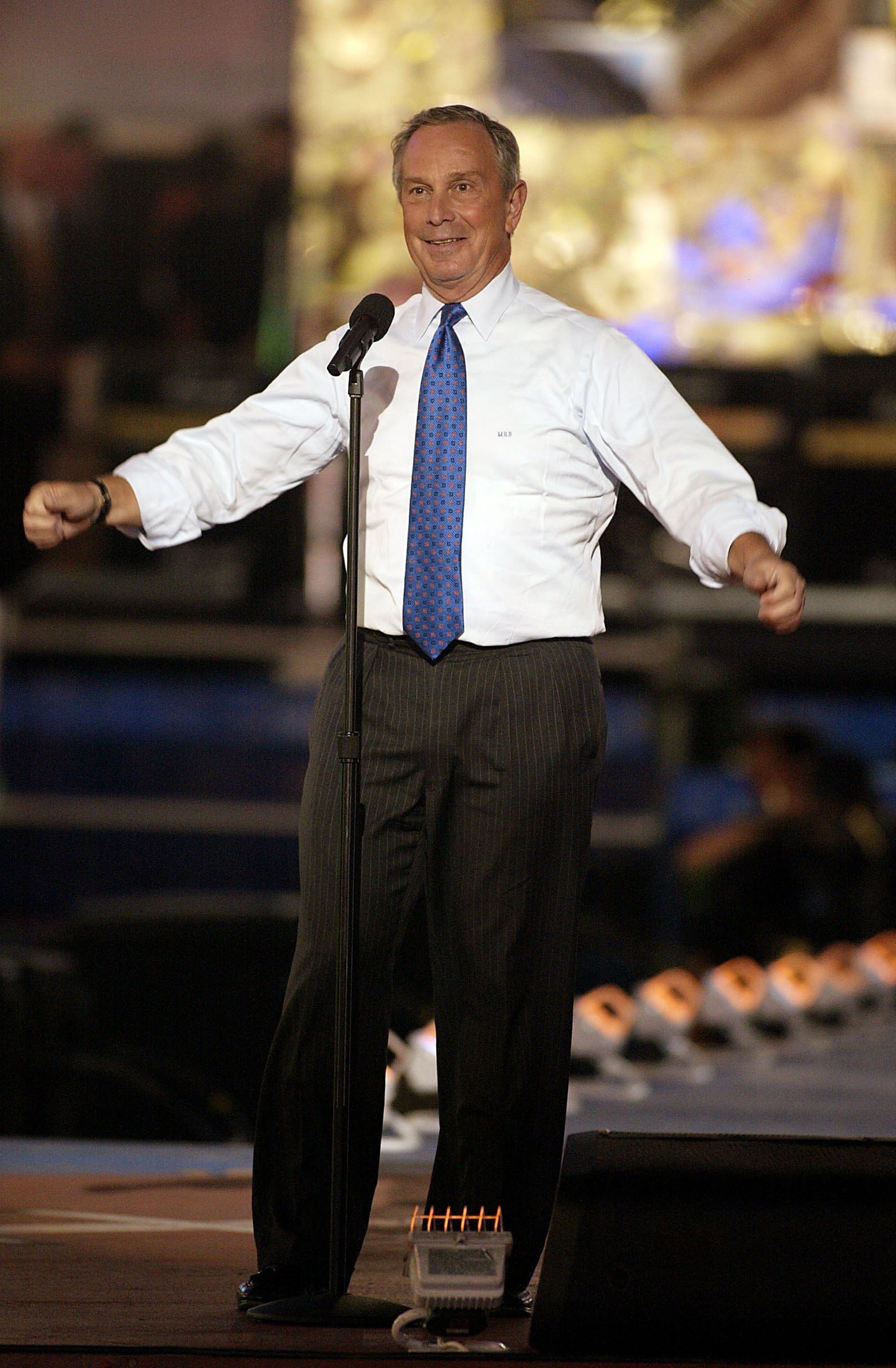 Person in a white shirt and tie speaks animatedly on stage, holding a microphone