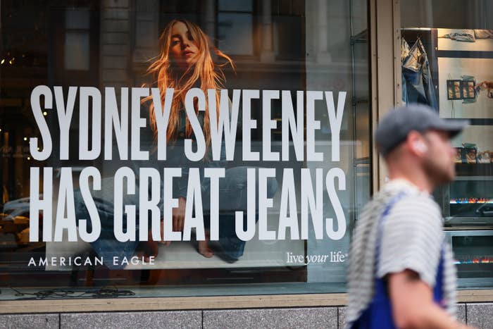 A store window showcases an ad reading "Sydney Sweeney Has Great Jeans" with her posing, while a person walks by outside