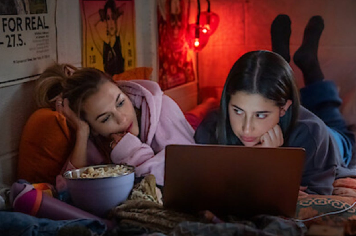 Two people lying on a bed, watching a laptop together. A bowl of popcorn is nearby. Cozy setting with posters and string lights on the wall