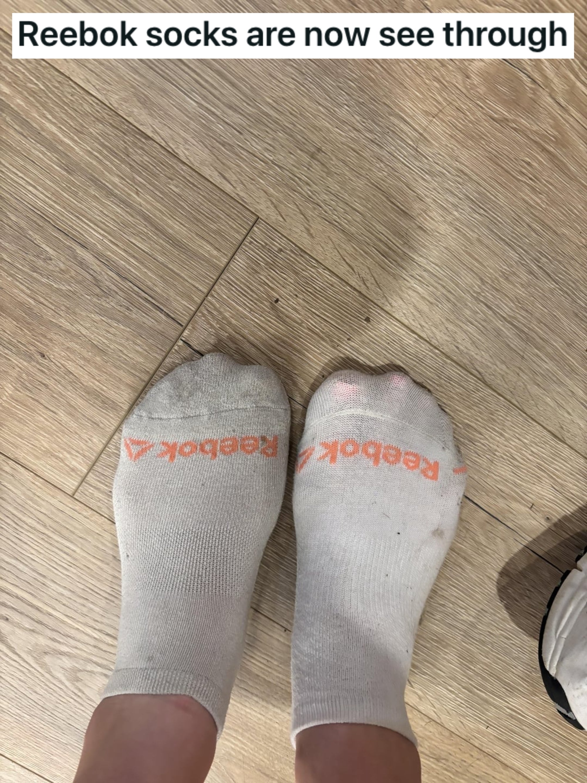 Two sock-covered feet connected  a woody  floor, wearing achromatic  Reebok socks