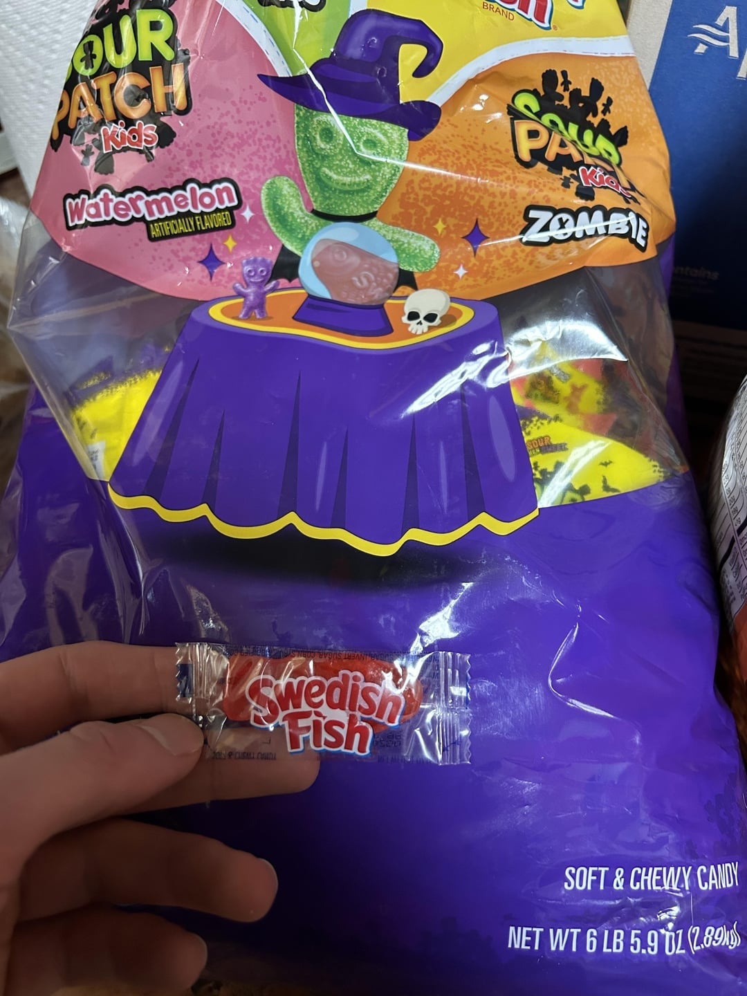 A manus  holds a Swedish Fish candy against a Halloween-themed candy container  with a cartoon ghoul and cauldron design