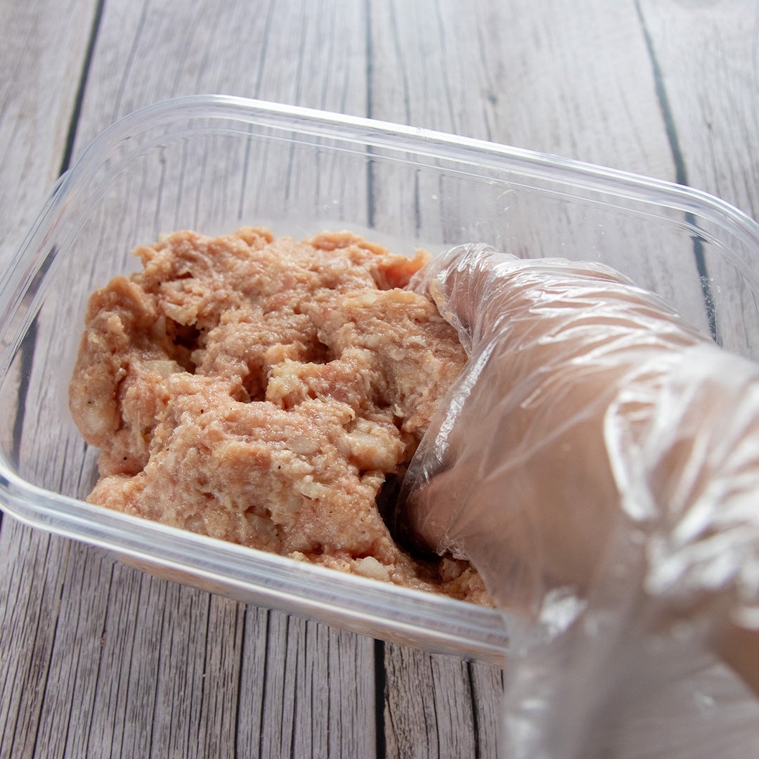 手袋をした手がプラスチック容器の中のひき肉を混ぜている様子。