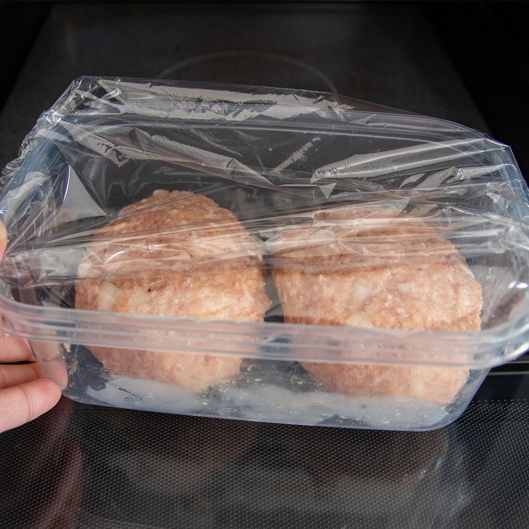 電子レンジの中でプラスチックの容器に入れられた2つのハンバーグのパティ。容器はラップで覆われている。