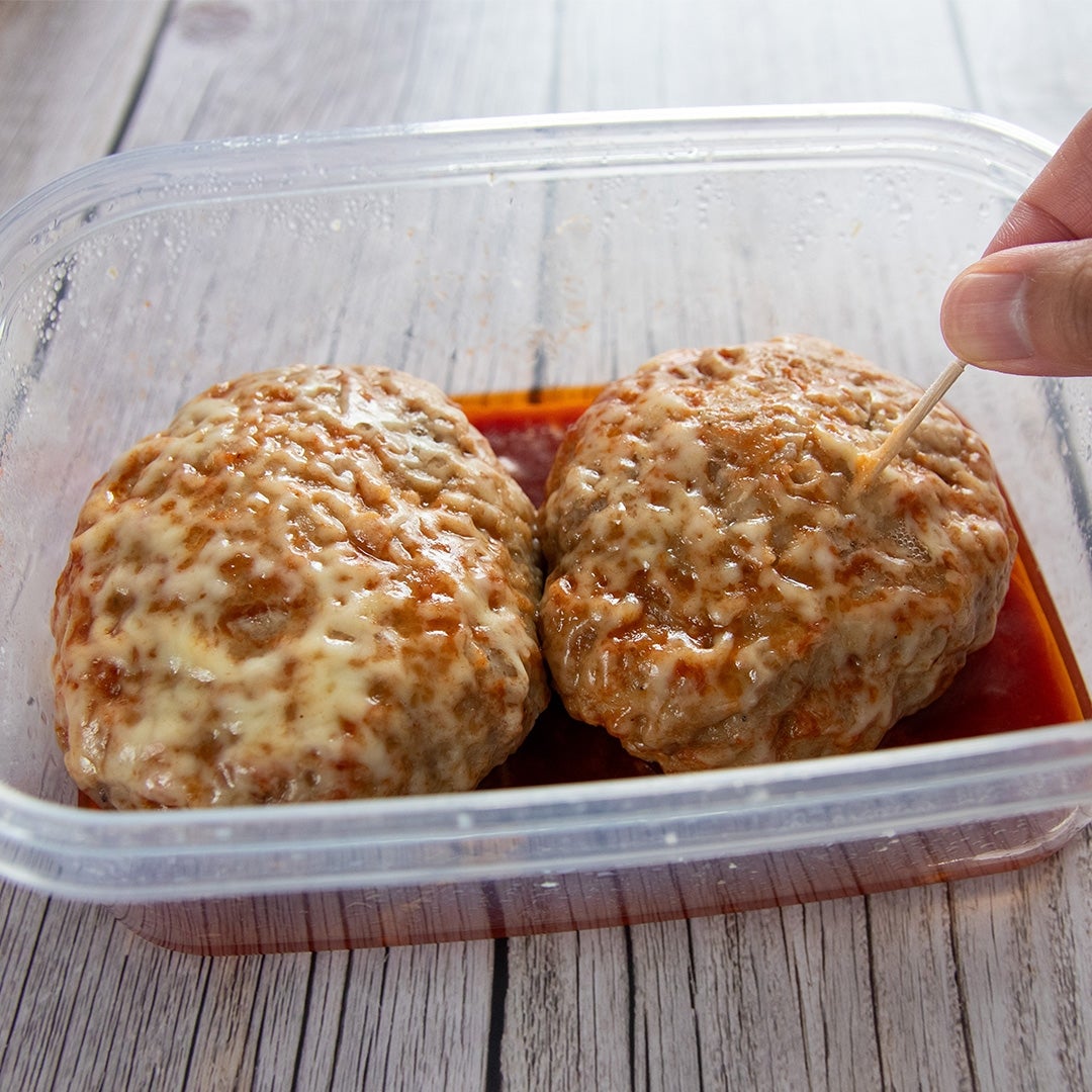 プラスチック容器に入ったチーズがのったハンバーグ2個。ソースが敷かれた状態で、つまようじでひとつを刺している。