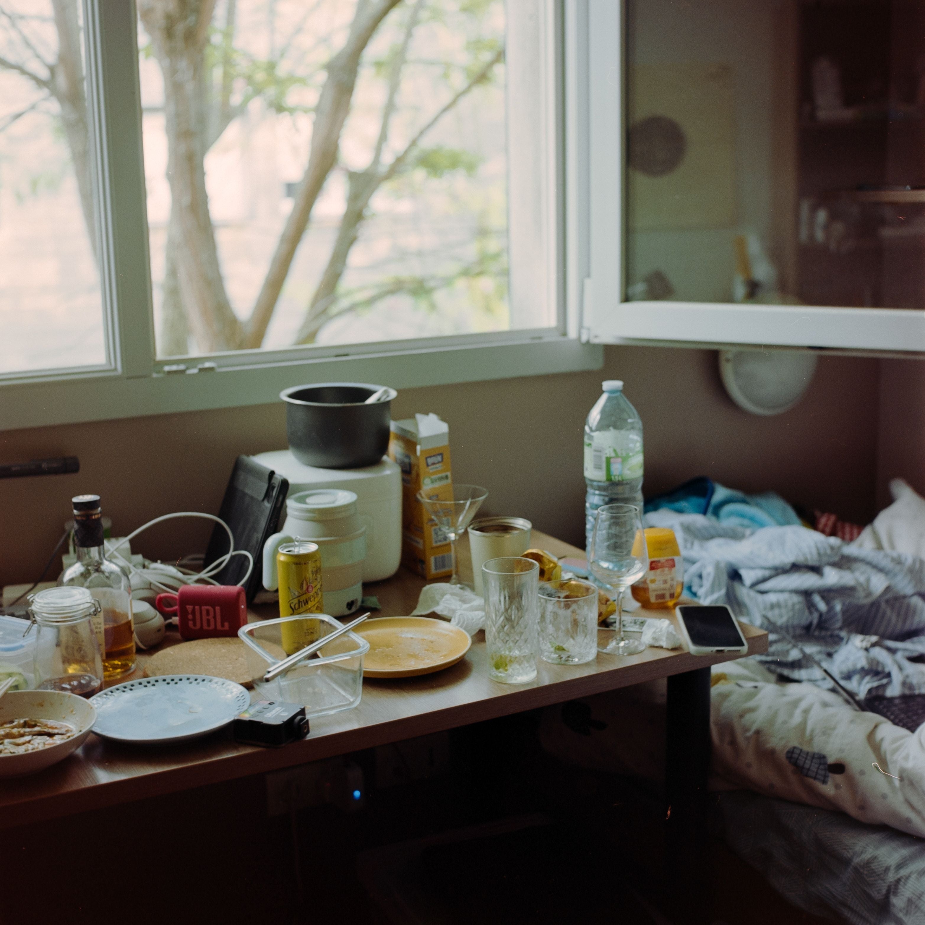 A cluttered desk with used dishes, beverages, and electronic items next to an unmade bed by a window