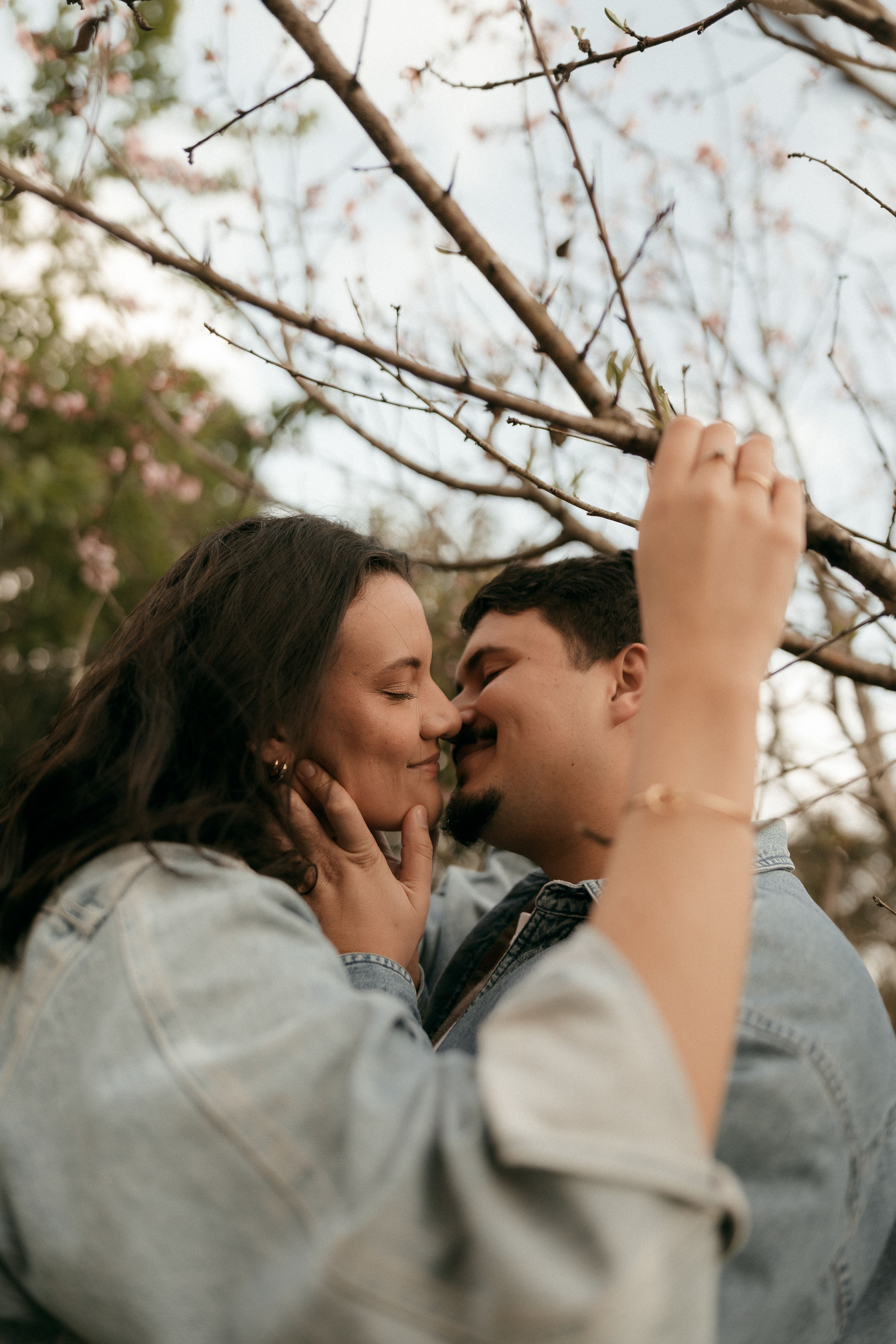 A couple in denim jackets shares a tender moment, embracing under a tree with delicate branches