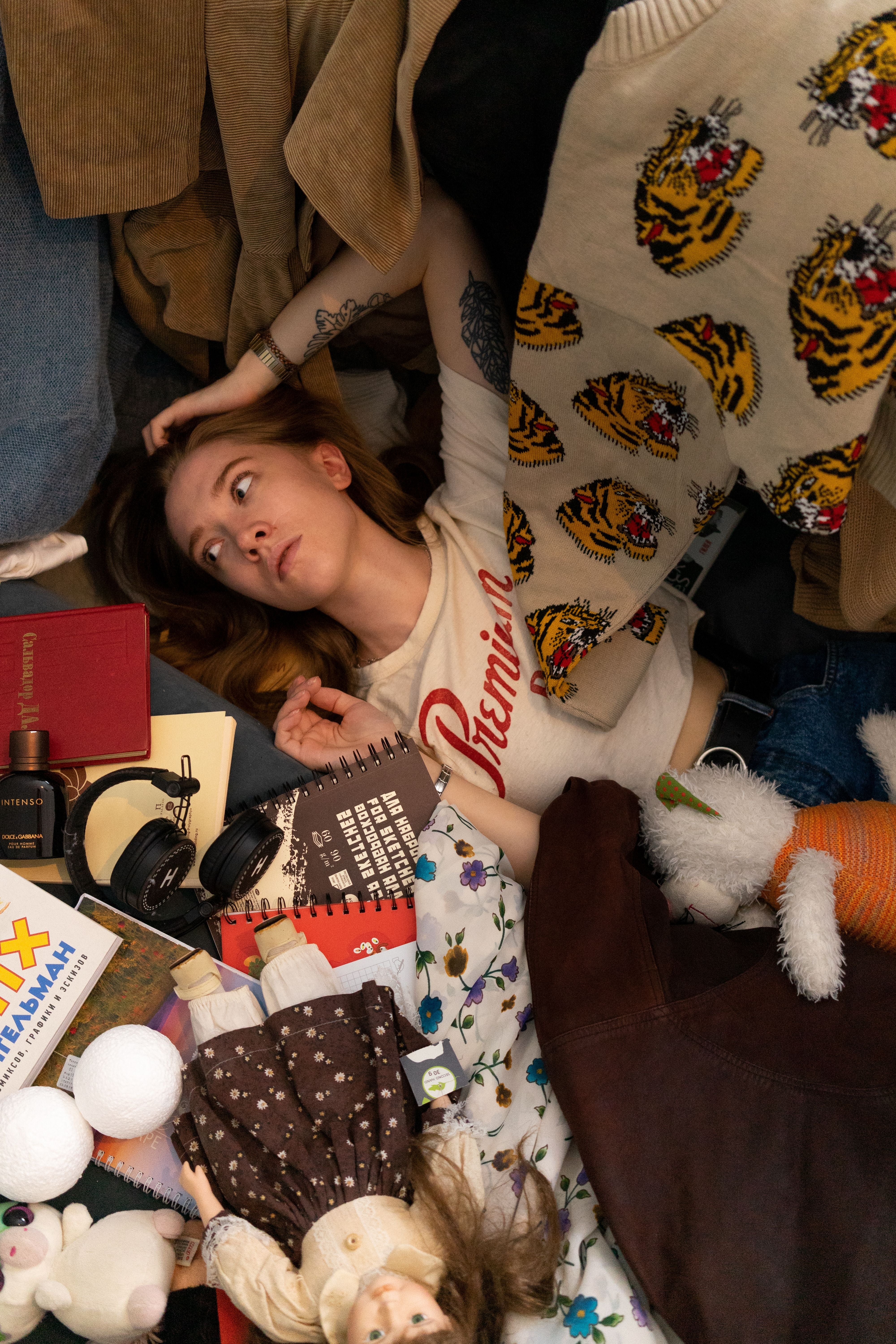 Woman lying among clothes, books, and toys, looking contemplative. She wears a T-shirt with a retro style print