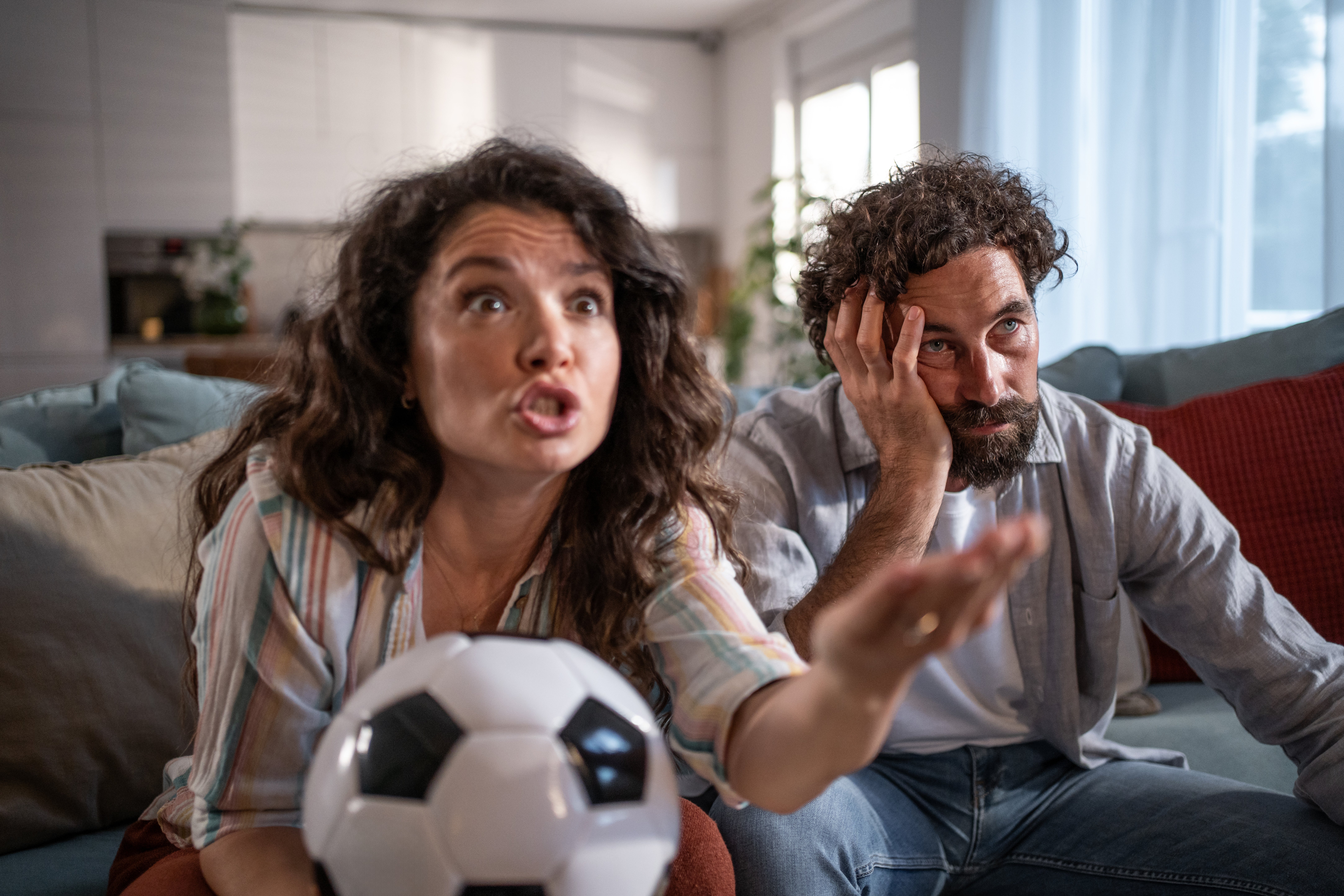 A man and woman sit on a couch watching TV. She looks animated with a soccer ball, while he appears tired or frustrated