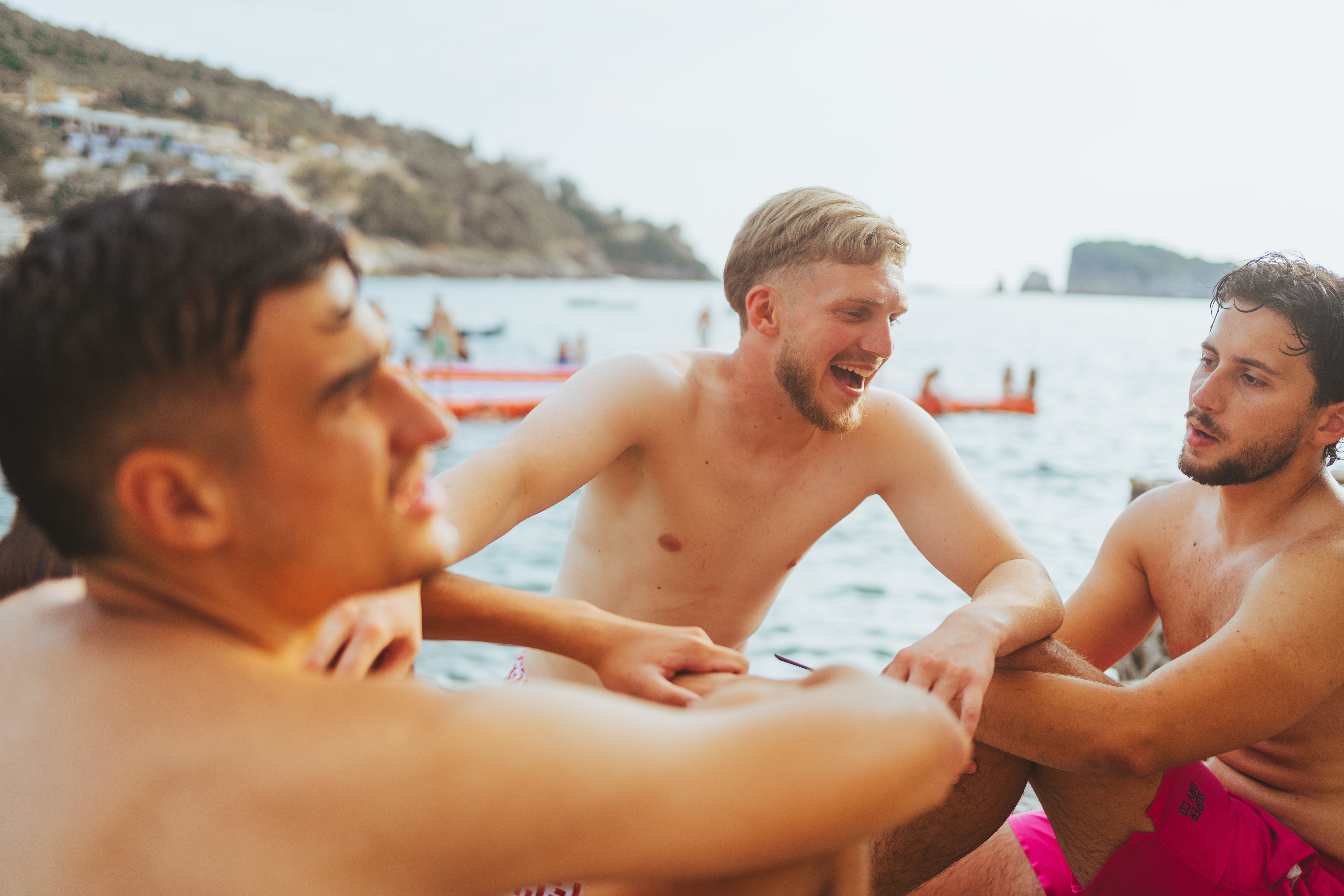 Three men in swim trunks sitting and talking by the sea, appearing relaxed and enjoying a sunny day