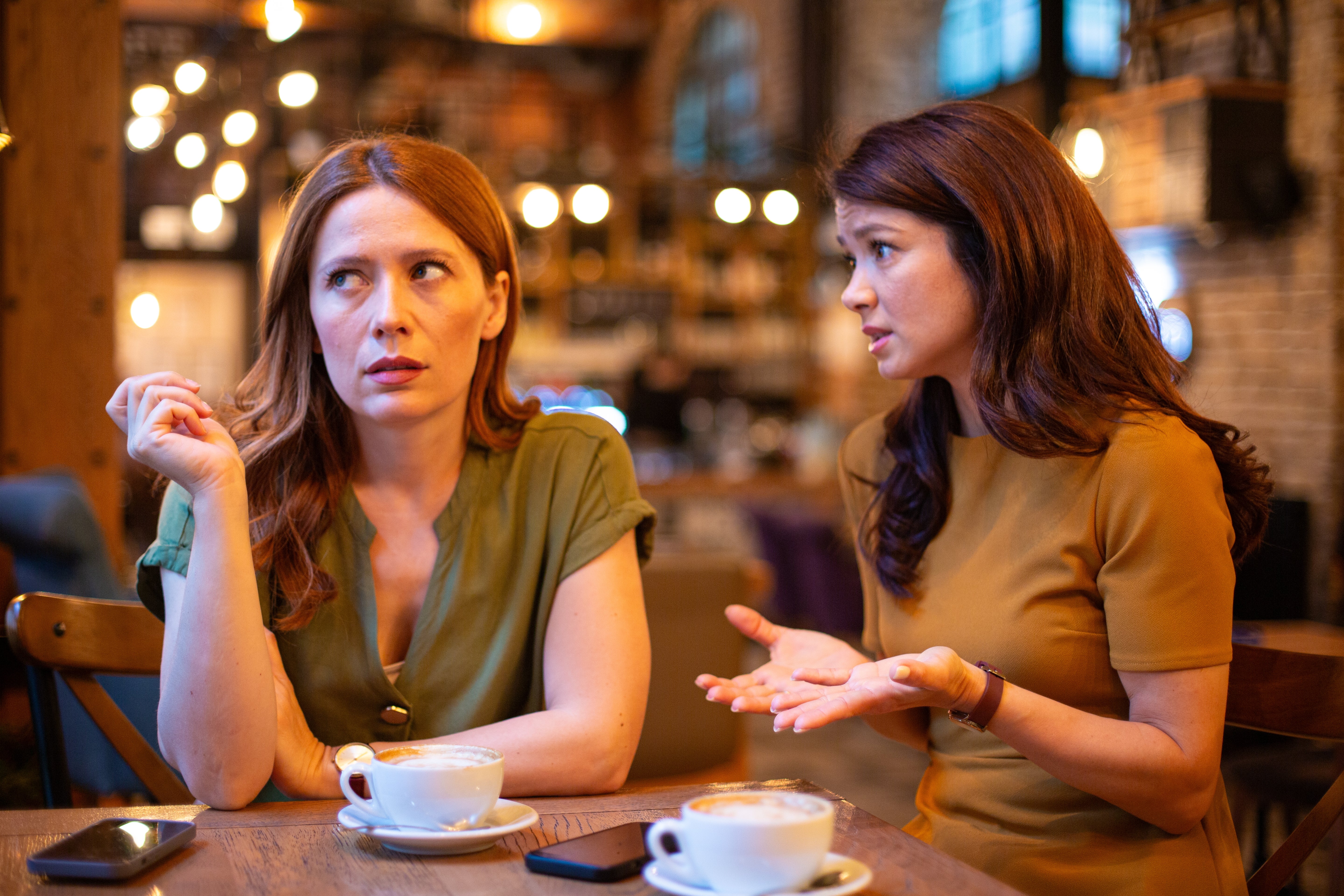 Two women sit in a cafe, engaged in a tense conversation. One looks frustrated while the other gestures as she talks