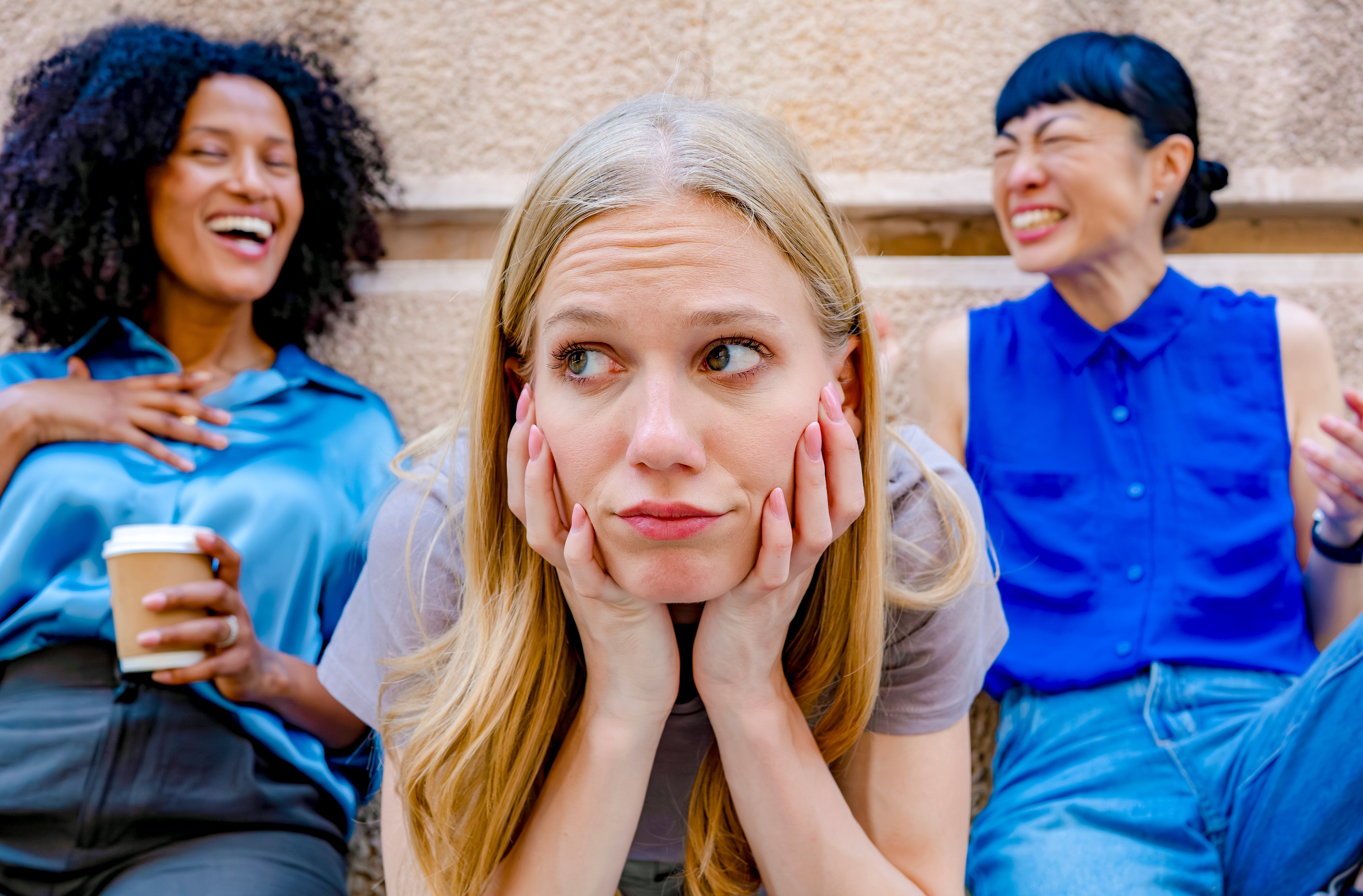 Three women sitting outdoors, two laughing, and one in the center looking concerned, resting her face on her hands. Casual attire
