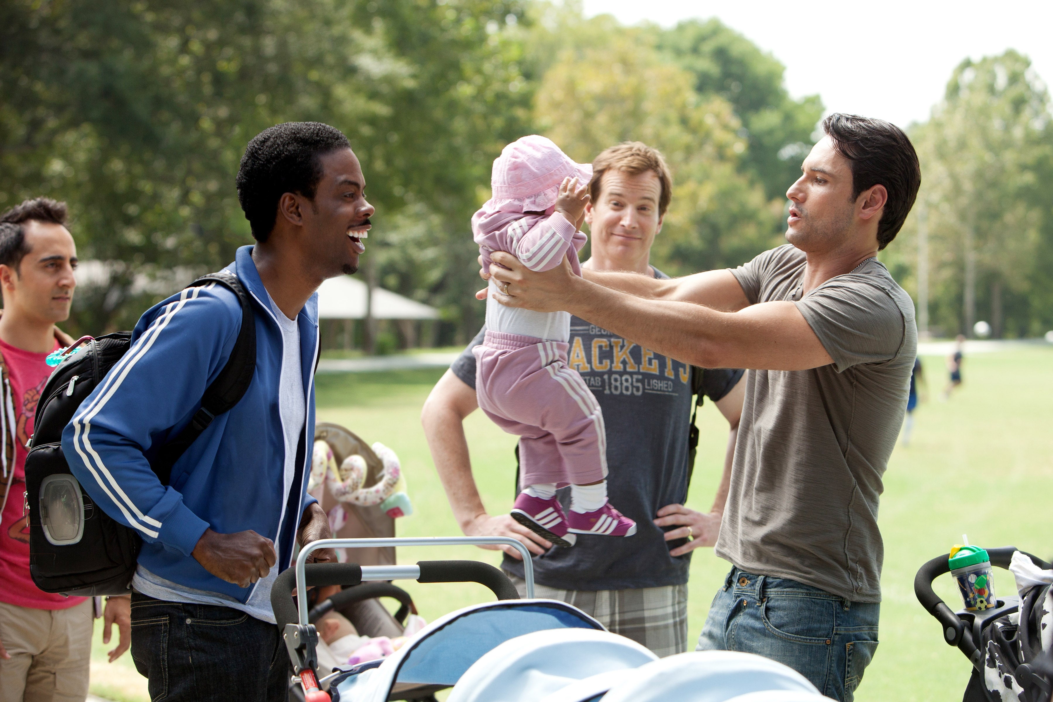 Group of friends in a park smiling as one man holds a baby, while others stand nearby with strollers, enjoying a day out