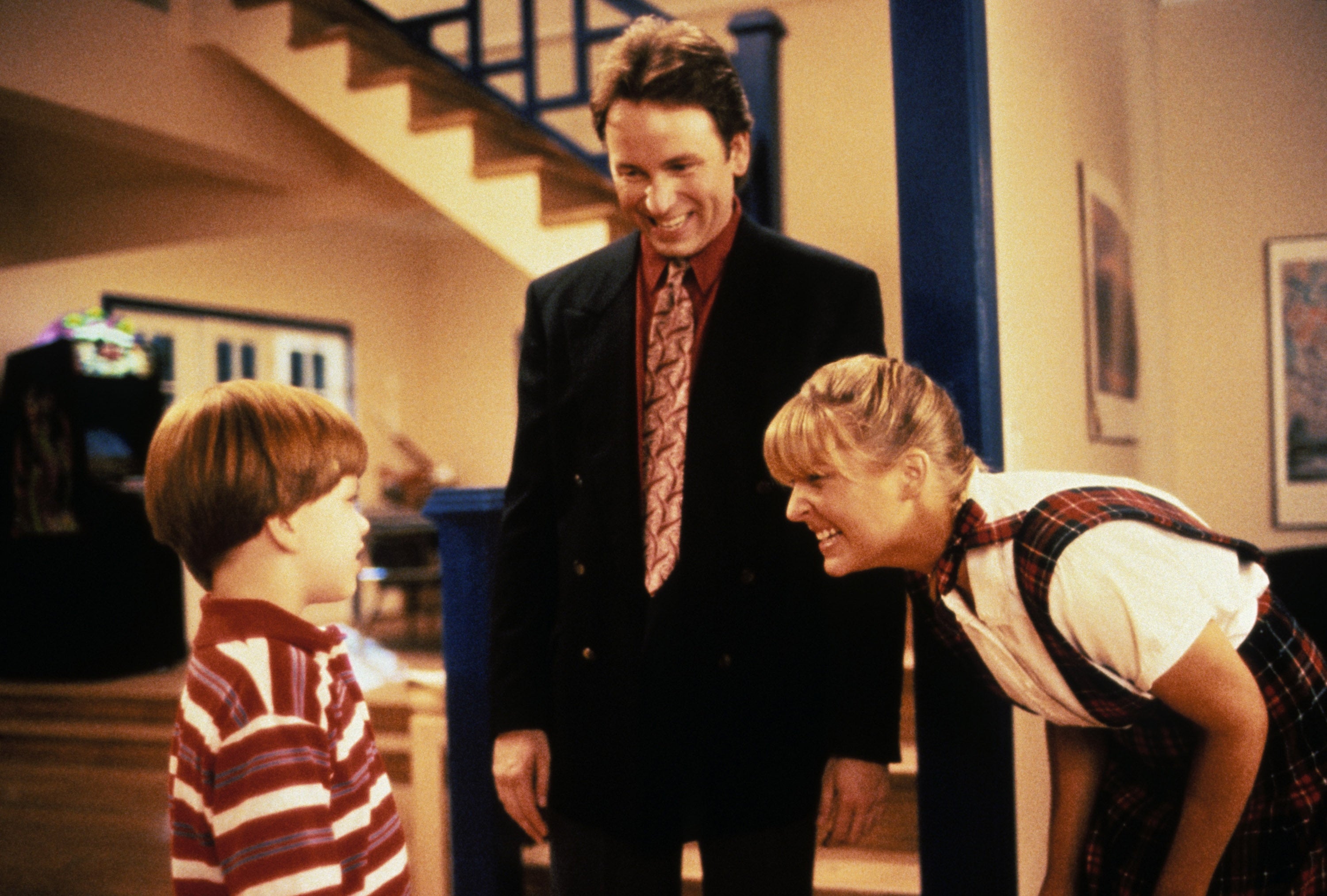 Child in front of two smiling adults in a home setting, one kneeling down. The mood is warm and family-oriented