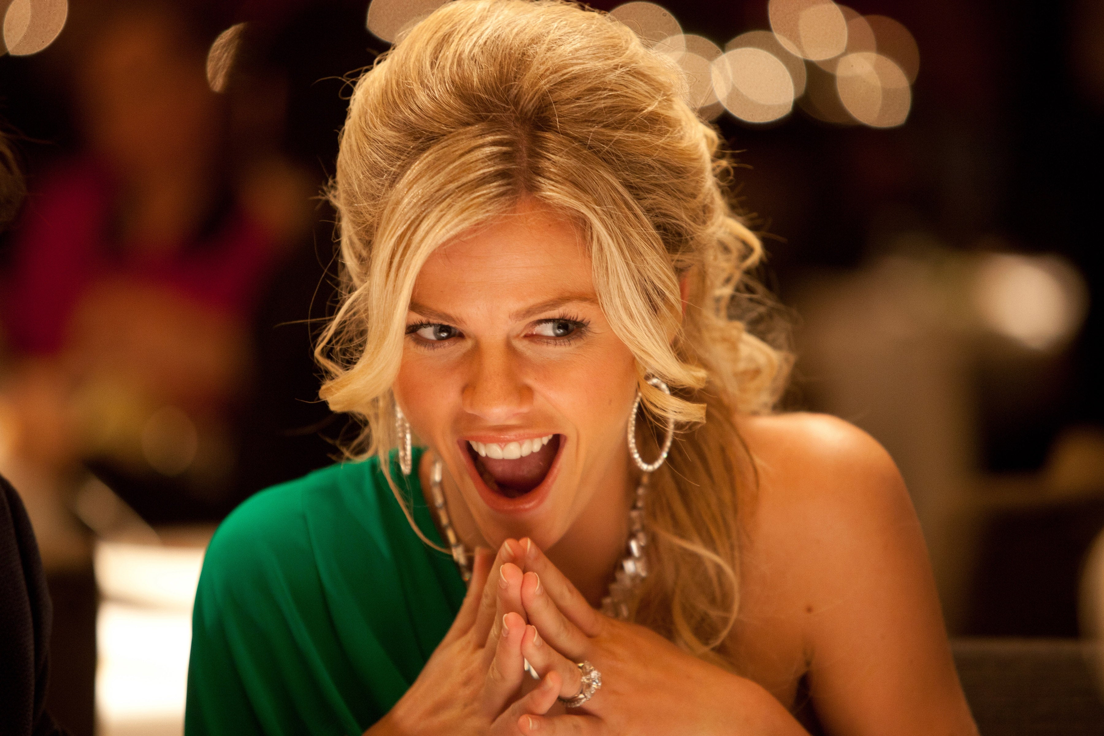 Woman with styled hair smiles and clasps hands at dinner. She wears a green dress and hoop earrings