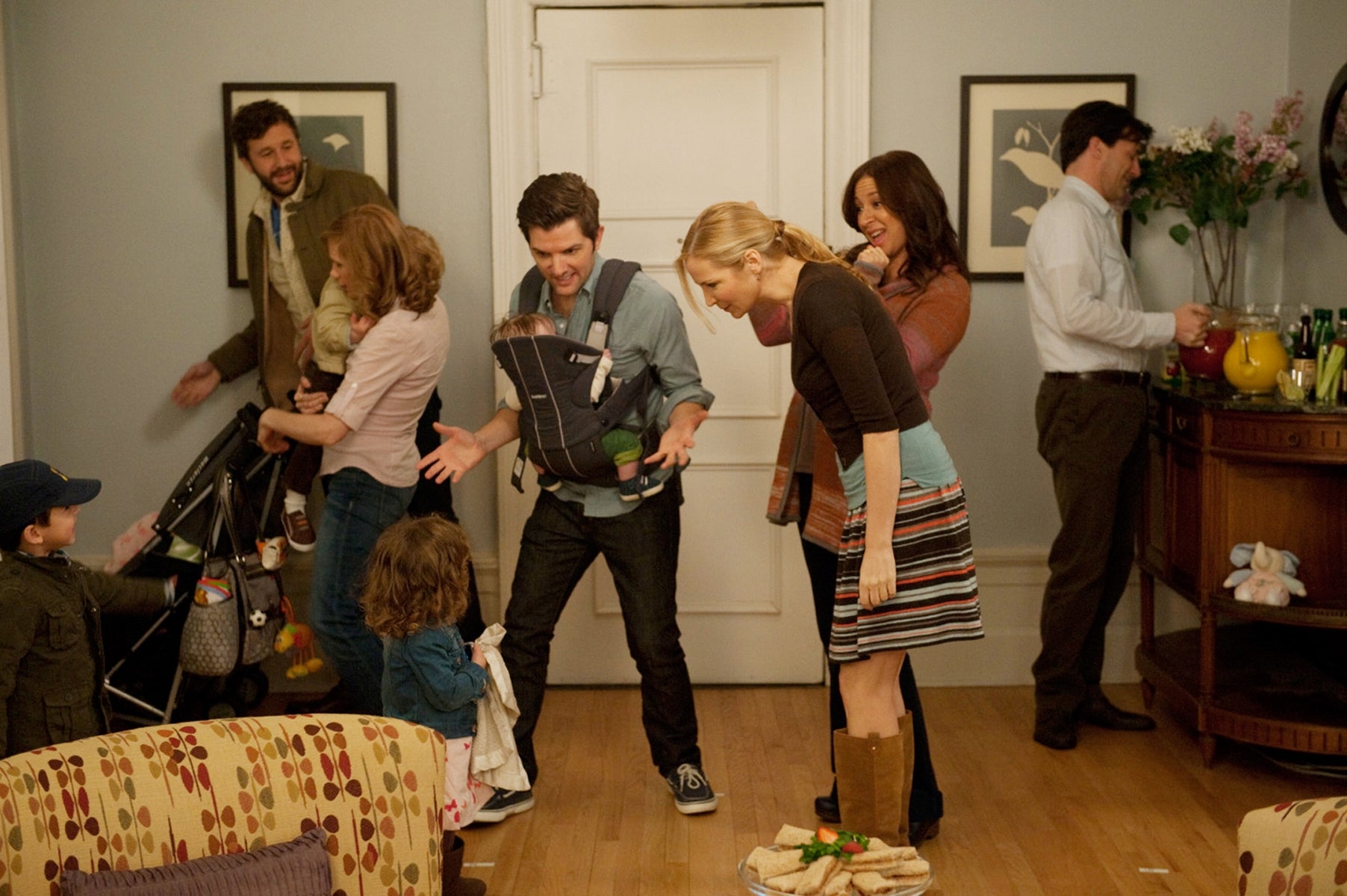 Group of adults and children in a home setting, interacting and talking, with casual clothing and warm expressions, suggests a family gathering