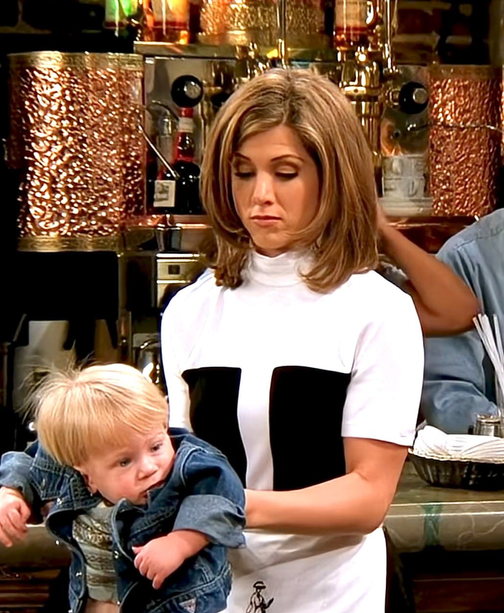 Person in a coffee shop holds a toddler, who wears a denim jacket. Person's style is trendy with a bold patterned top