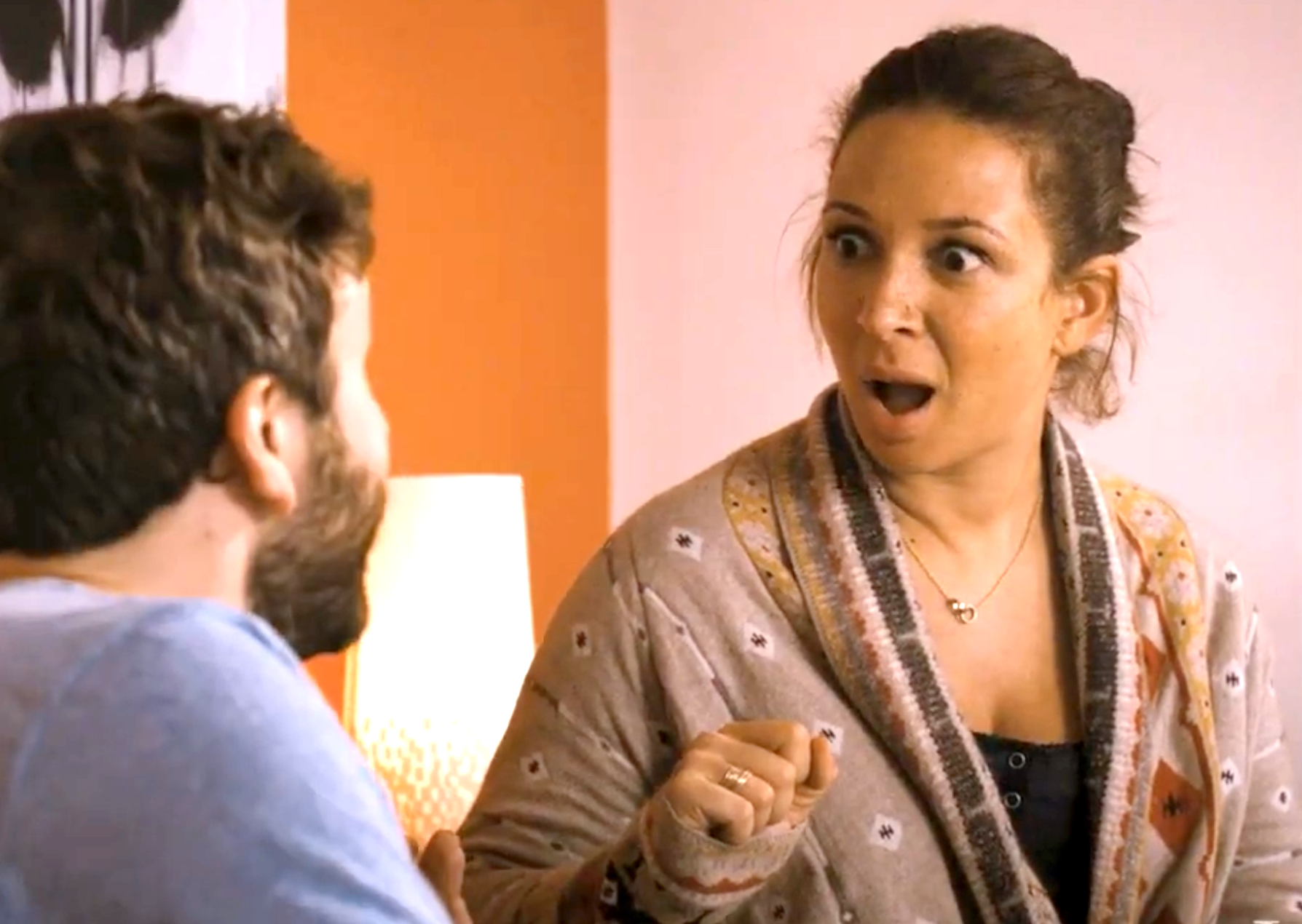 A surprised woman looks at a man, gesturing with an open mouth and raised eyebrows, in a casual setting at home