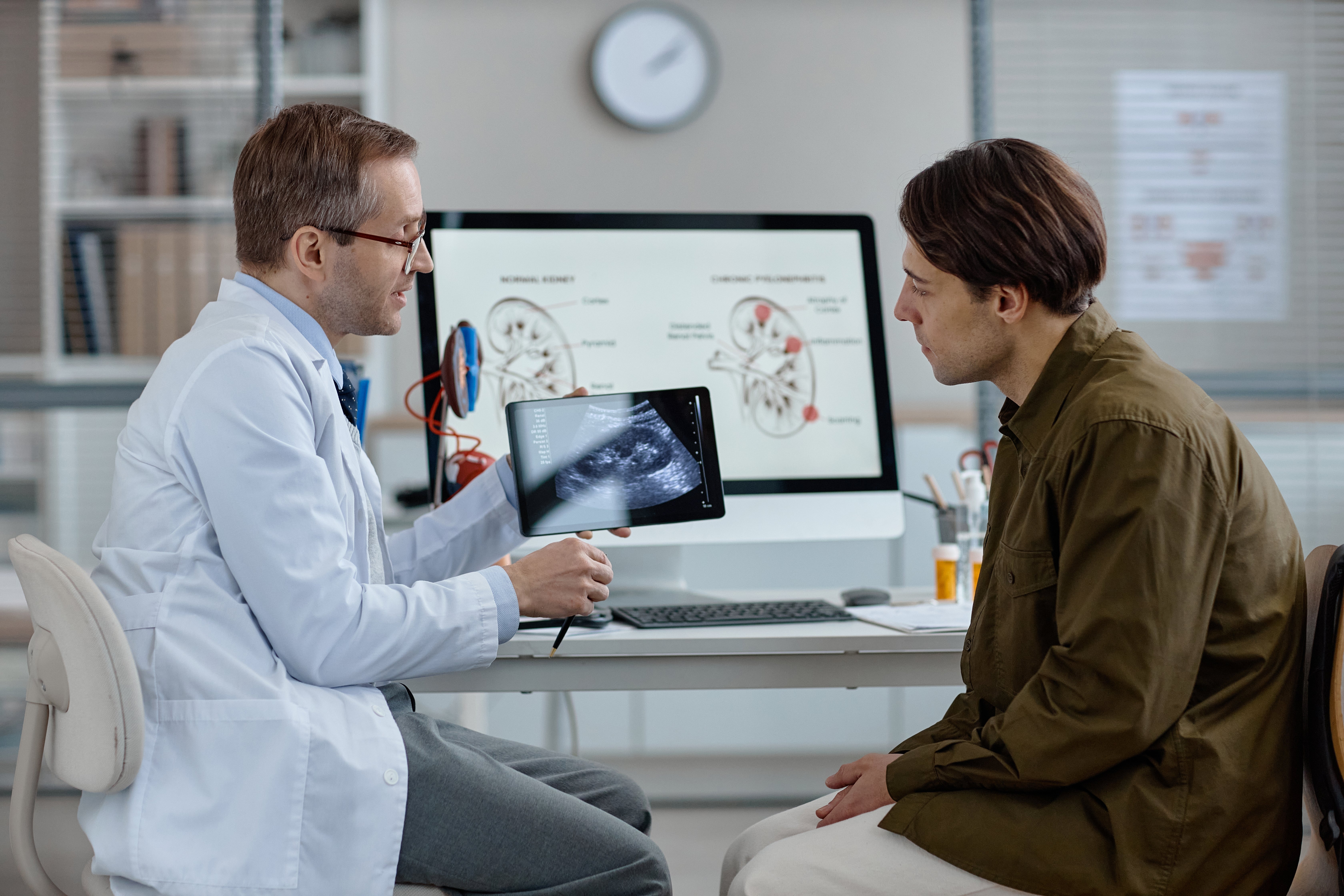 Doctor showing a patient a tablet displaying a medical scan in a consultation room with anatomical charts in the background