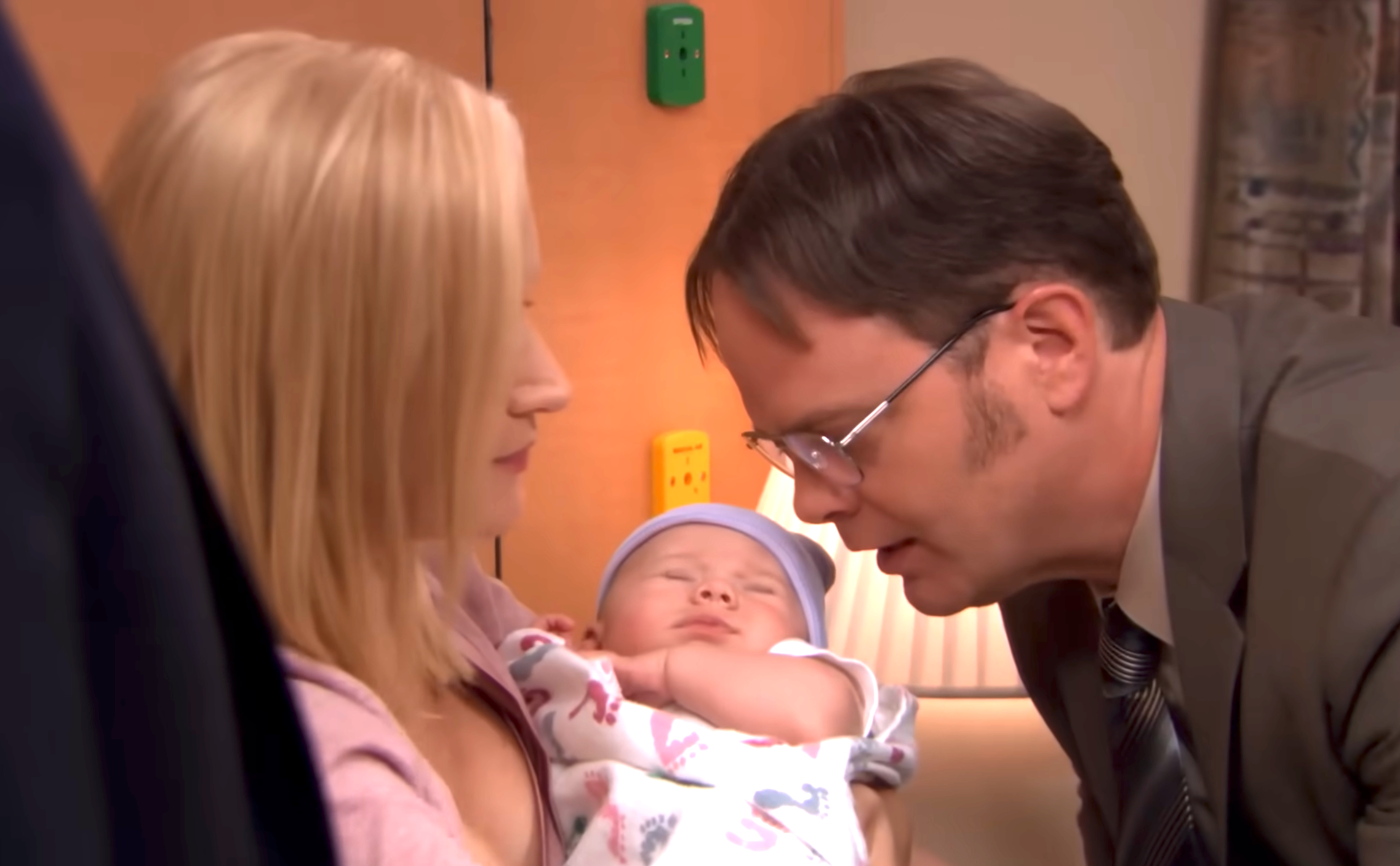 A woman holds a sleeping baby while a man in glasses leans in, speaking gently to the infant