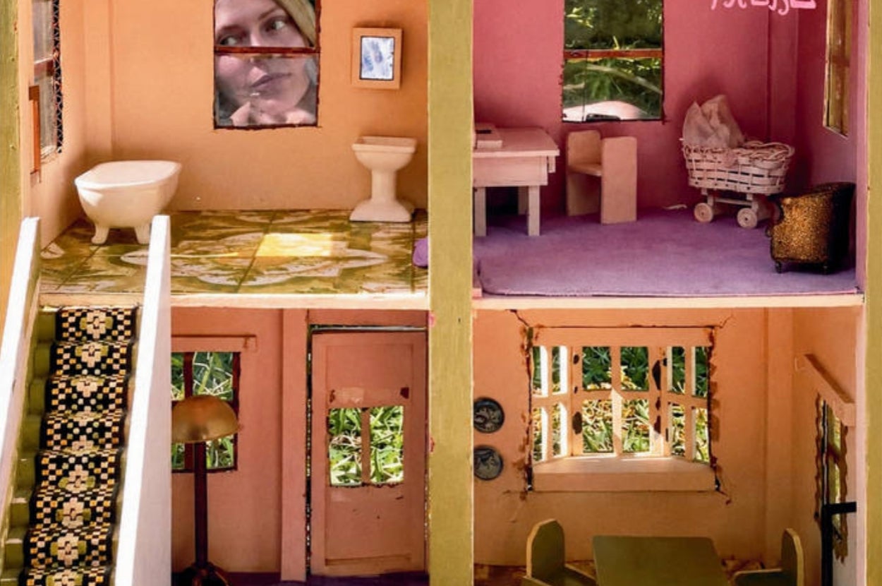A person peers through a window of a dollhouse with furniture visible inside, including a bathroom, living area, and baby carriage