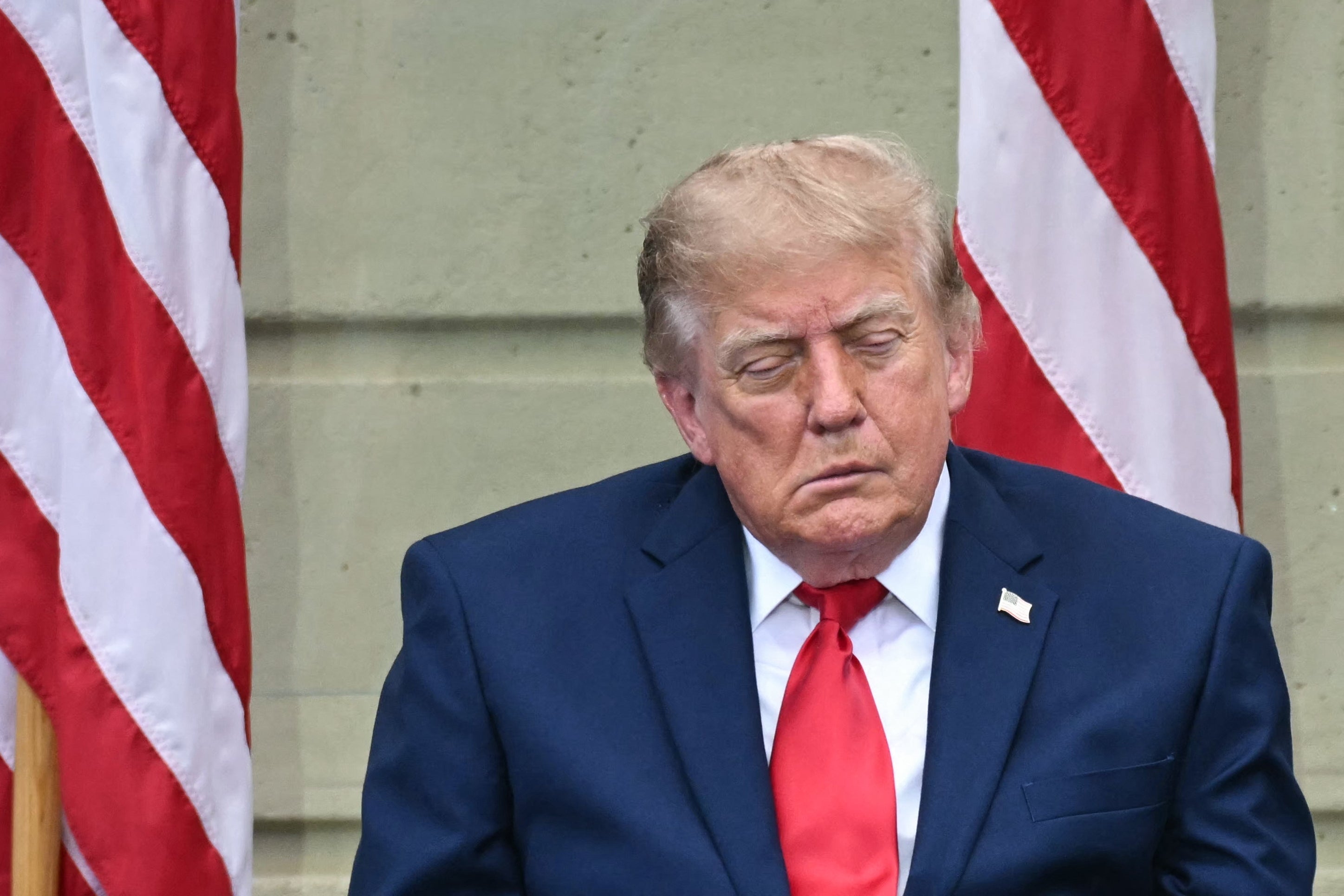 Donald Trump i dress med lukkede øyne, sittende mellom to amerikanske flagg