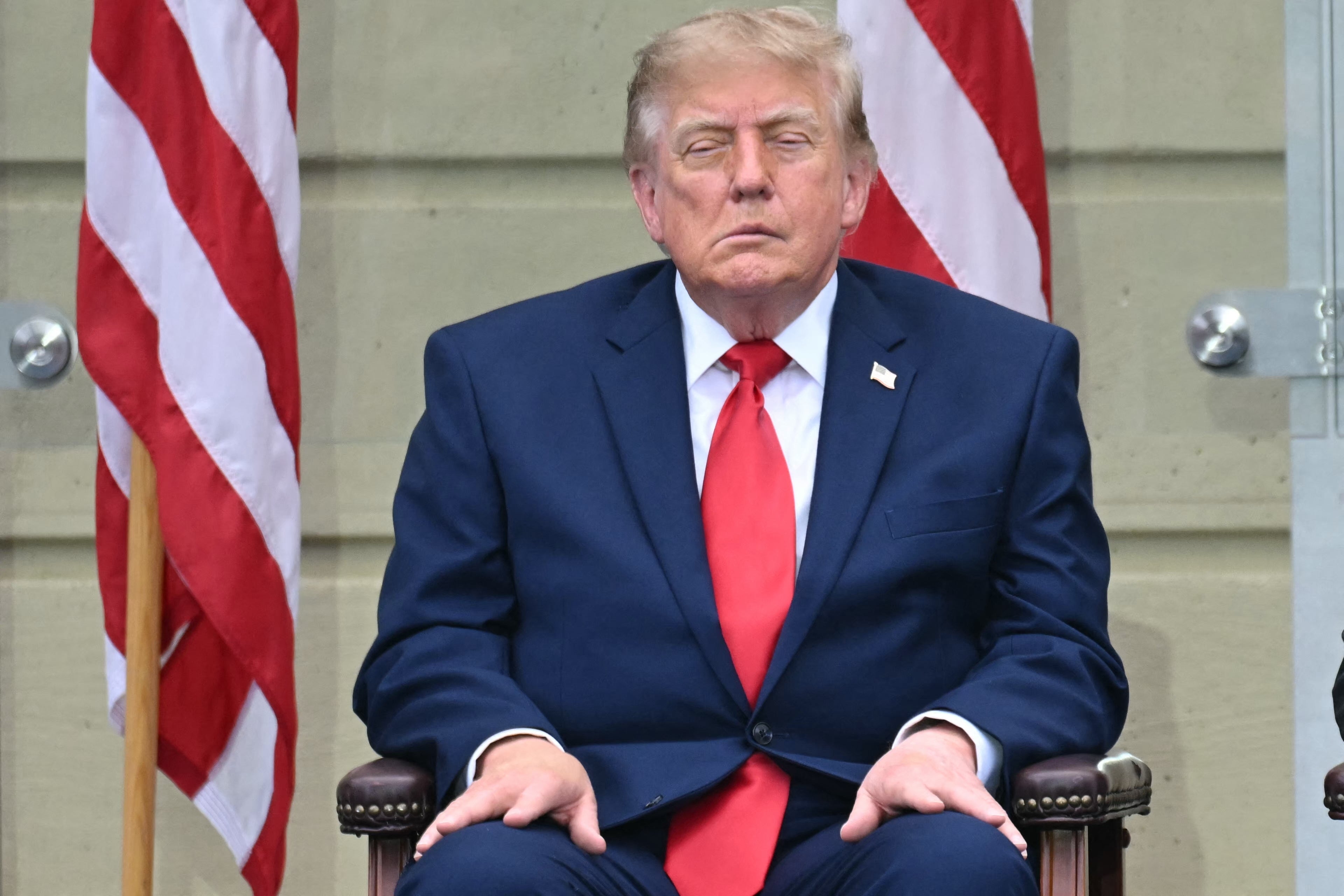 Donald Trump i blå dress og rødt slips sitter på en stol med synlige amerikanske flagg i bakgrunnen; høyre side av ansiktet er synlig hengende