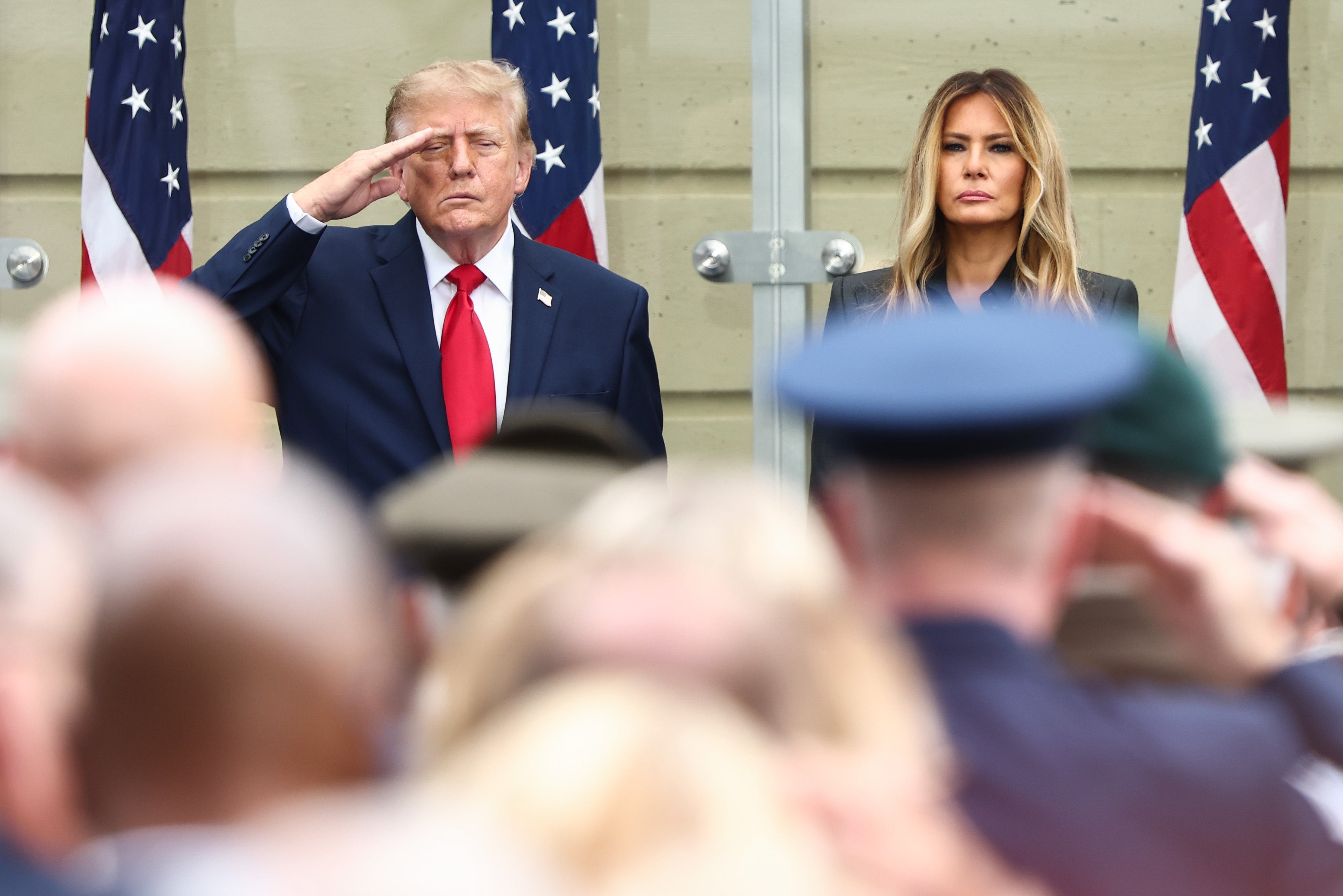 Donald Trump, i dress, hilser ved et formelt arrangement med flagg bak seg, stående ved siden av Melania i en lang frakk; nedre høyre munnvik er synlig hengende