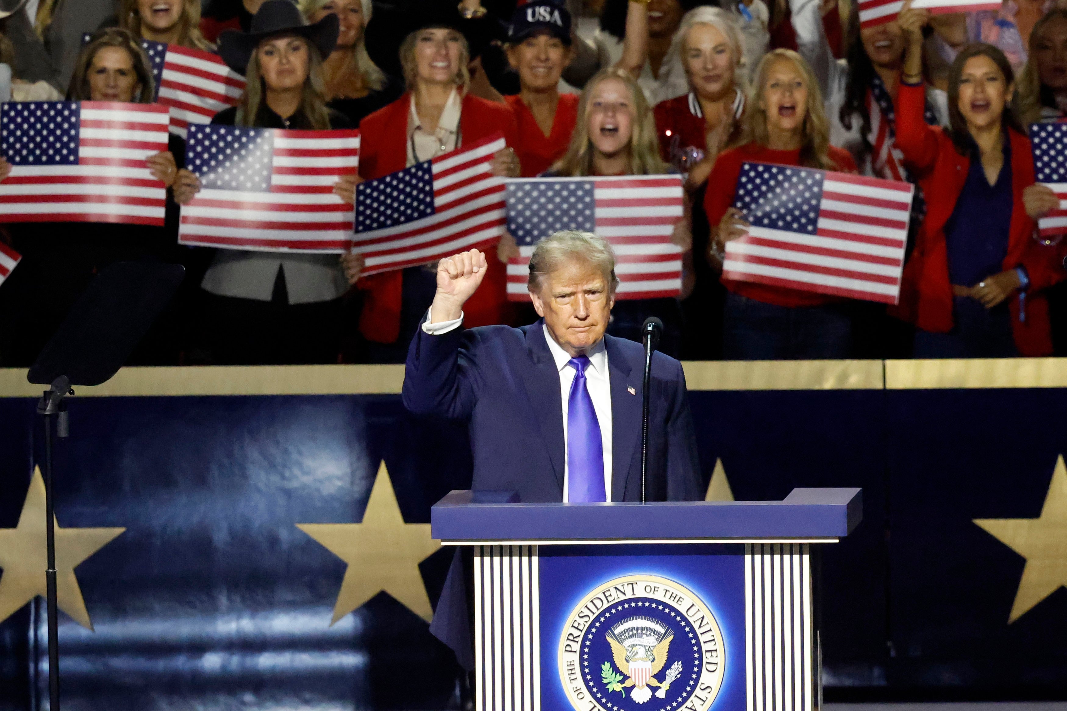 En person på et podium med en presidentsegl hever en knyttneve. Publikumsmedlemmer bak holder amerikanske flagg, og ser ut til å være på et offentlig arrangement eller rally