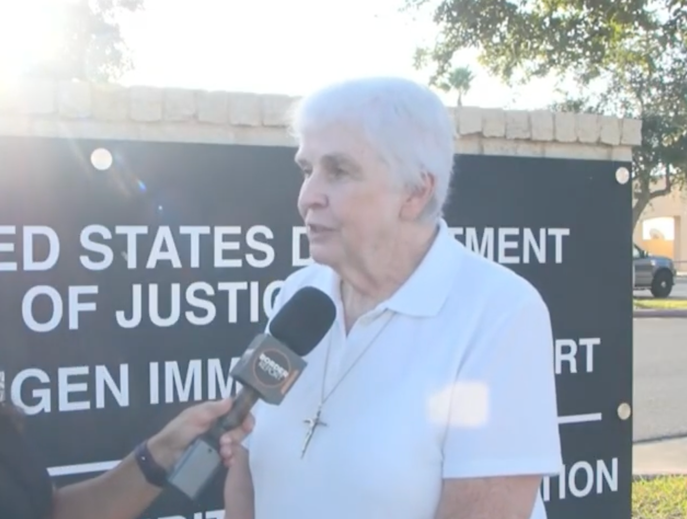 En eldre person som har på seg en hvit skjorte og et halskjede snakker inn i en mikrofon holdt av en intervjuer foran et skilt fra det amerikanske justisdepartementet
