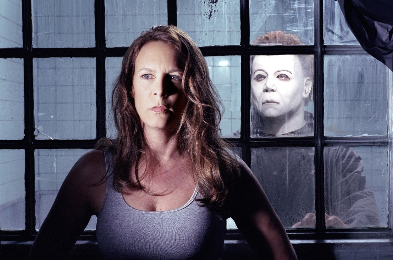 Jamie Lee Curtis in tank top stands in front of a window with a masked Michael Myers ominously behind her in "Halloween" scene
