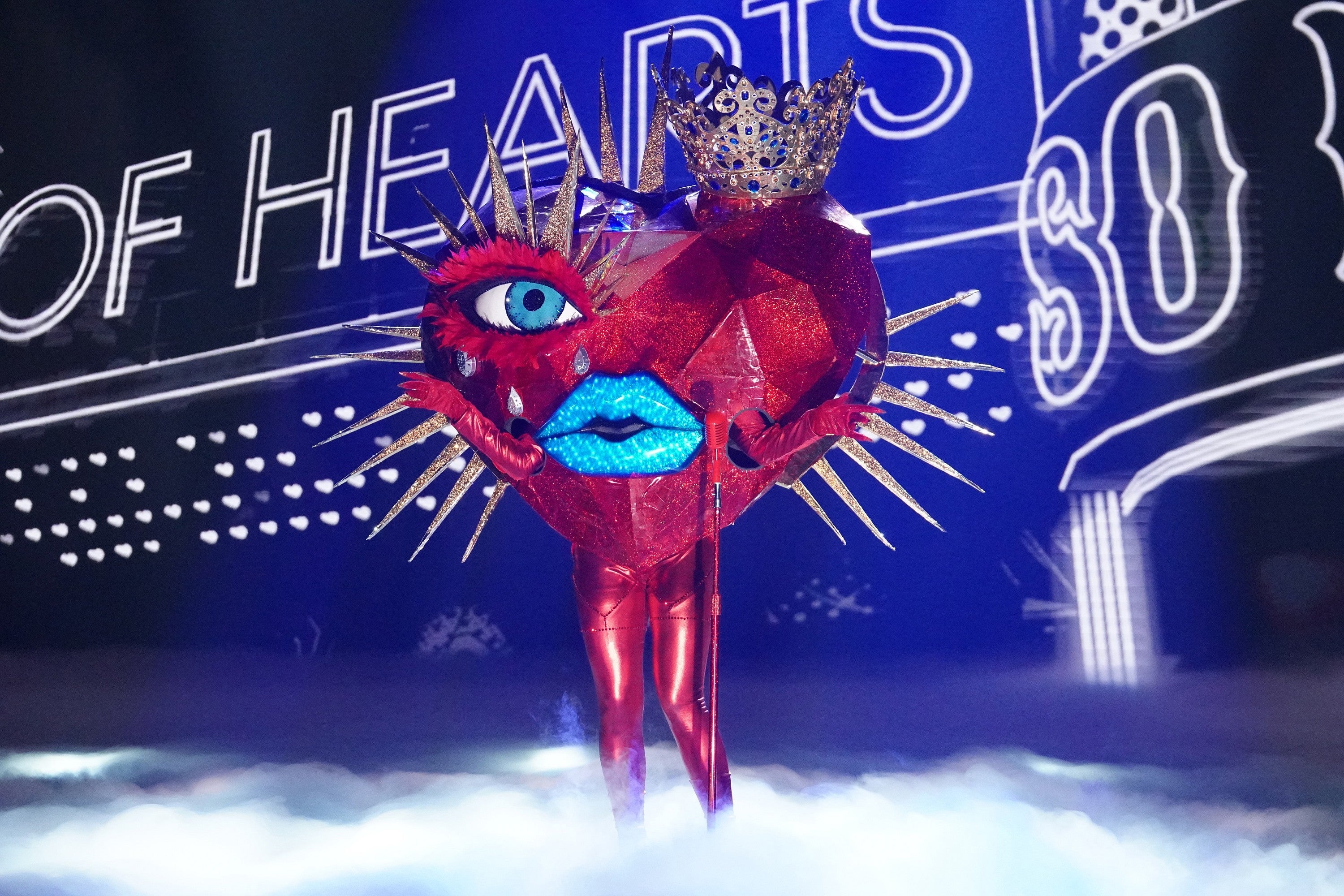 Person i et kostyme med rødt hjertetema med pigger, store øyne og lepper og utsmykket krone for The Masked Singer