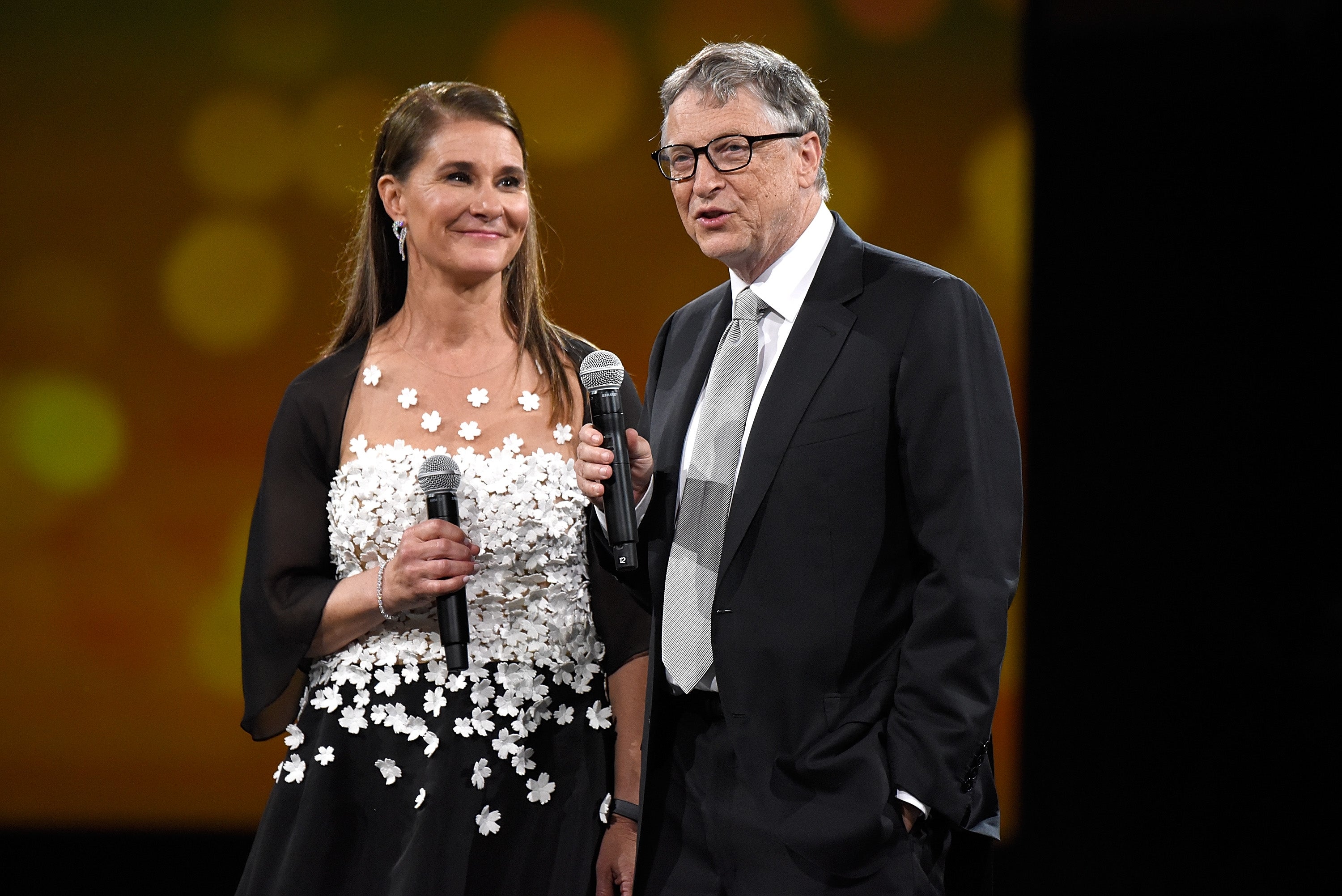Melinda French Gates and Bill Gates stand on stage in formal attire, holding microphones and smiling at an event. The woman's dress has floral accents