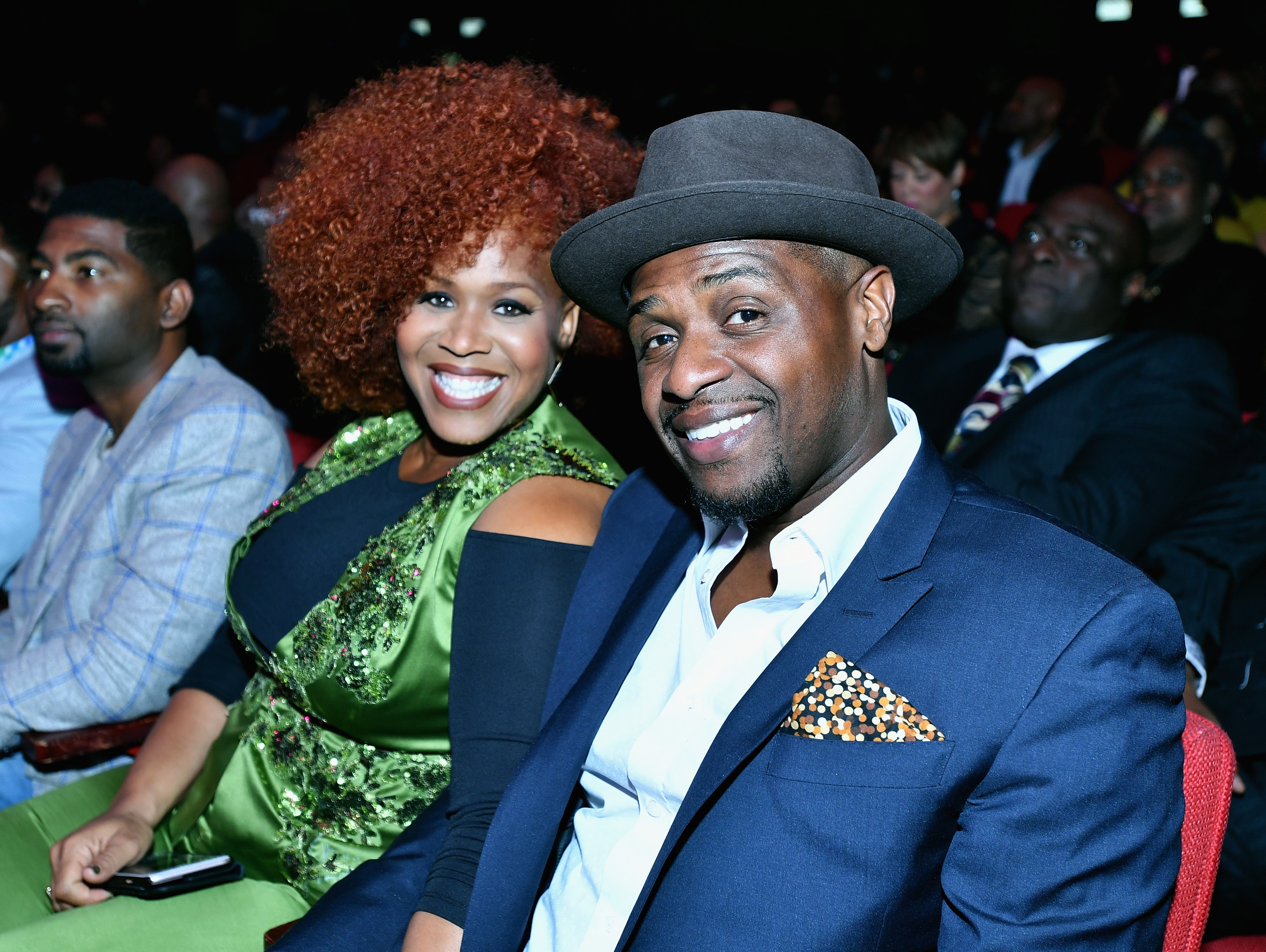 Tina Campbell and Teddy Campbell sit beside each other in an audience at an event. Tina wears a sequined green gown and Teddy wears a blue suit jacket and hat