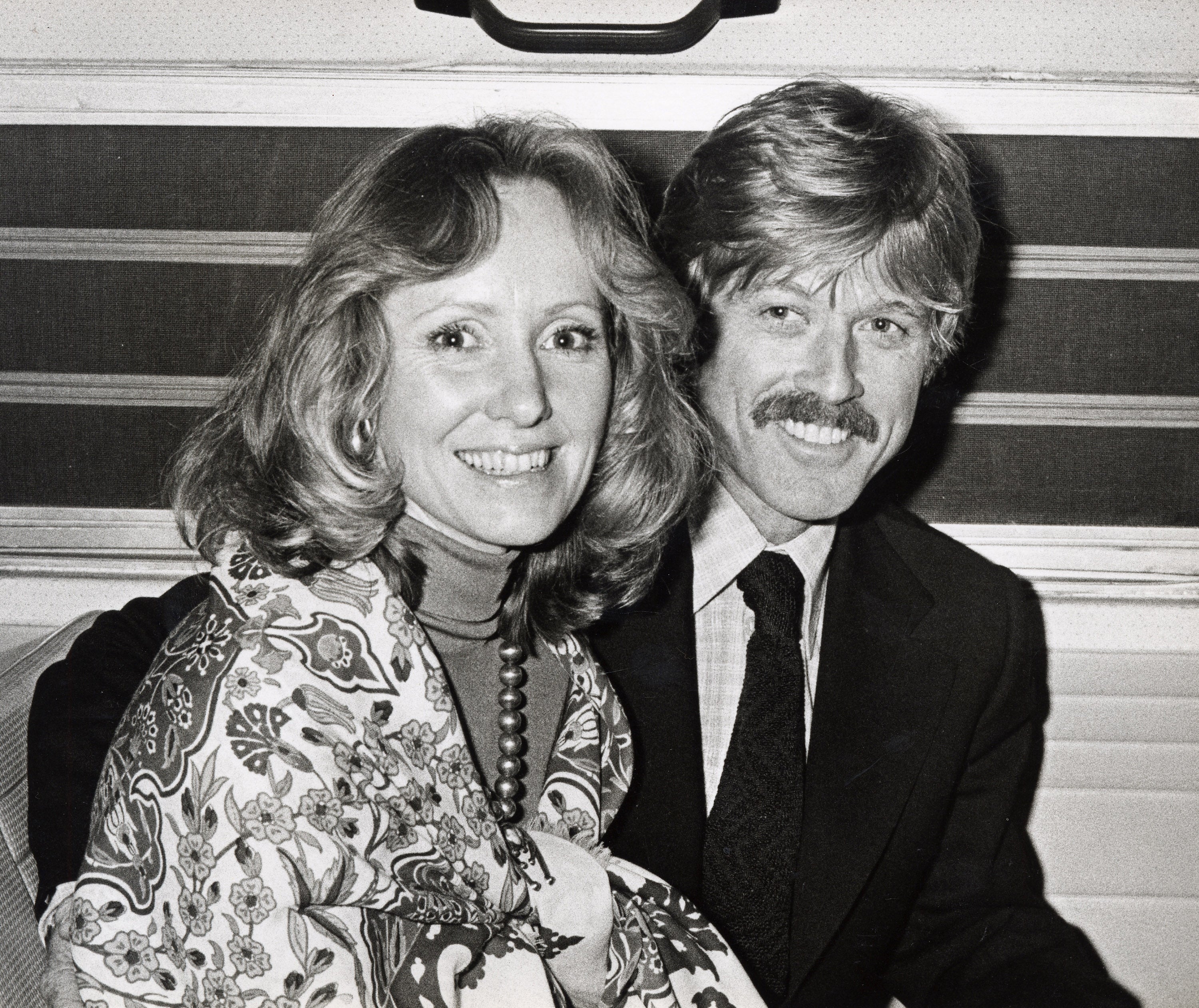 Lola Van Wagenen and Robert Redford smile as they sit. Van Wagenen wears a patterned coat and turtleneck, and Redford wears a suit and tie