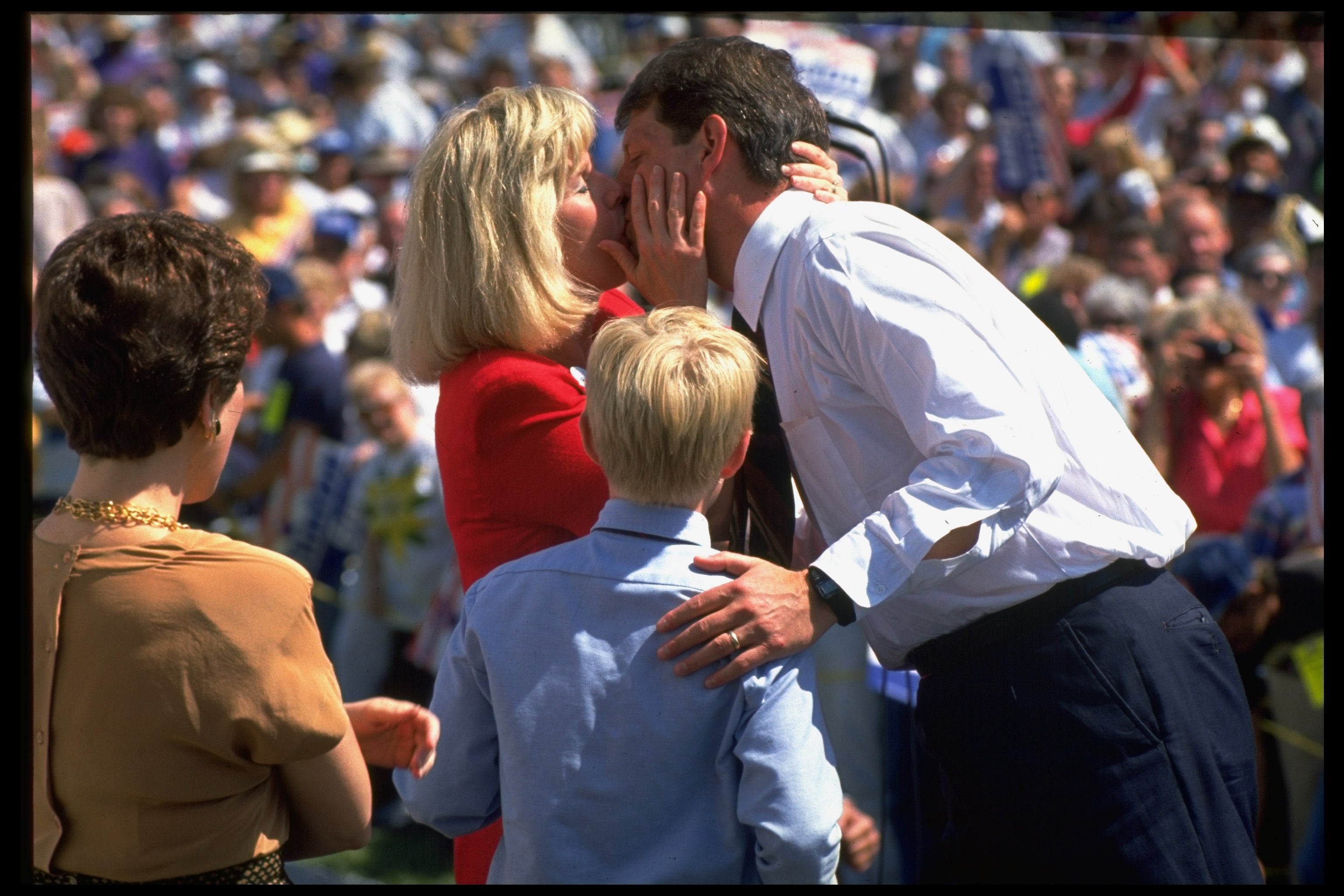 Al Gore and Tipper Gore share a kiss in a crowd at a campaign rally. A child stands nearby. Tipper wears a red blazer, and Al wears a button-up shirt and tie
