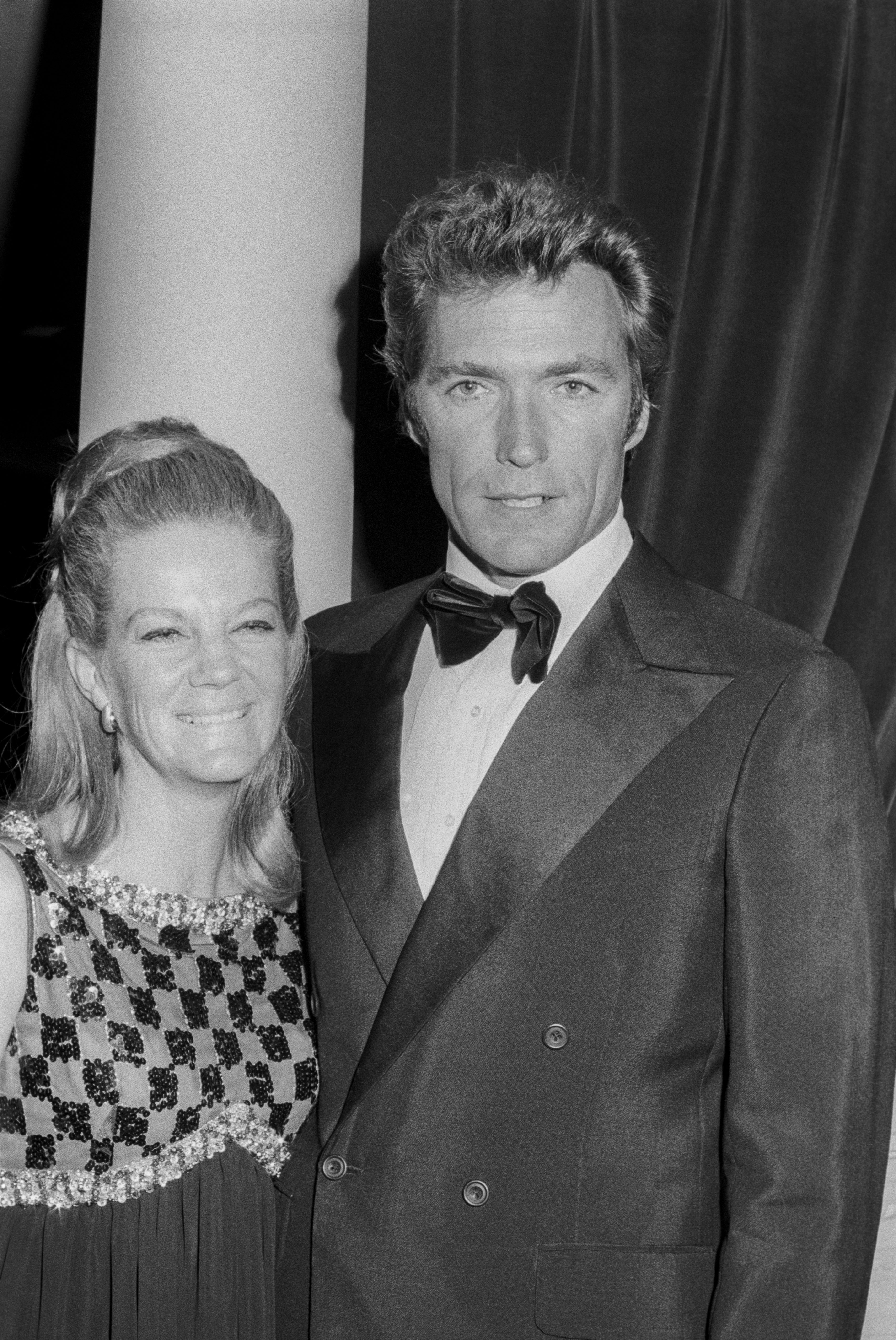 Maggie Johnson and Clint Eastwood pose together at an event. Johnson wears a patterned dress, and Eastwood wears a classic tuxedo