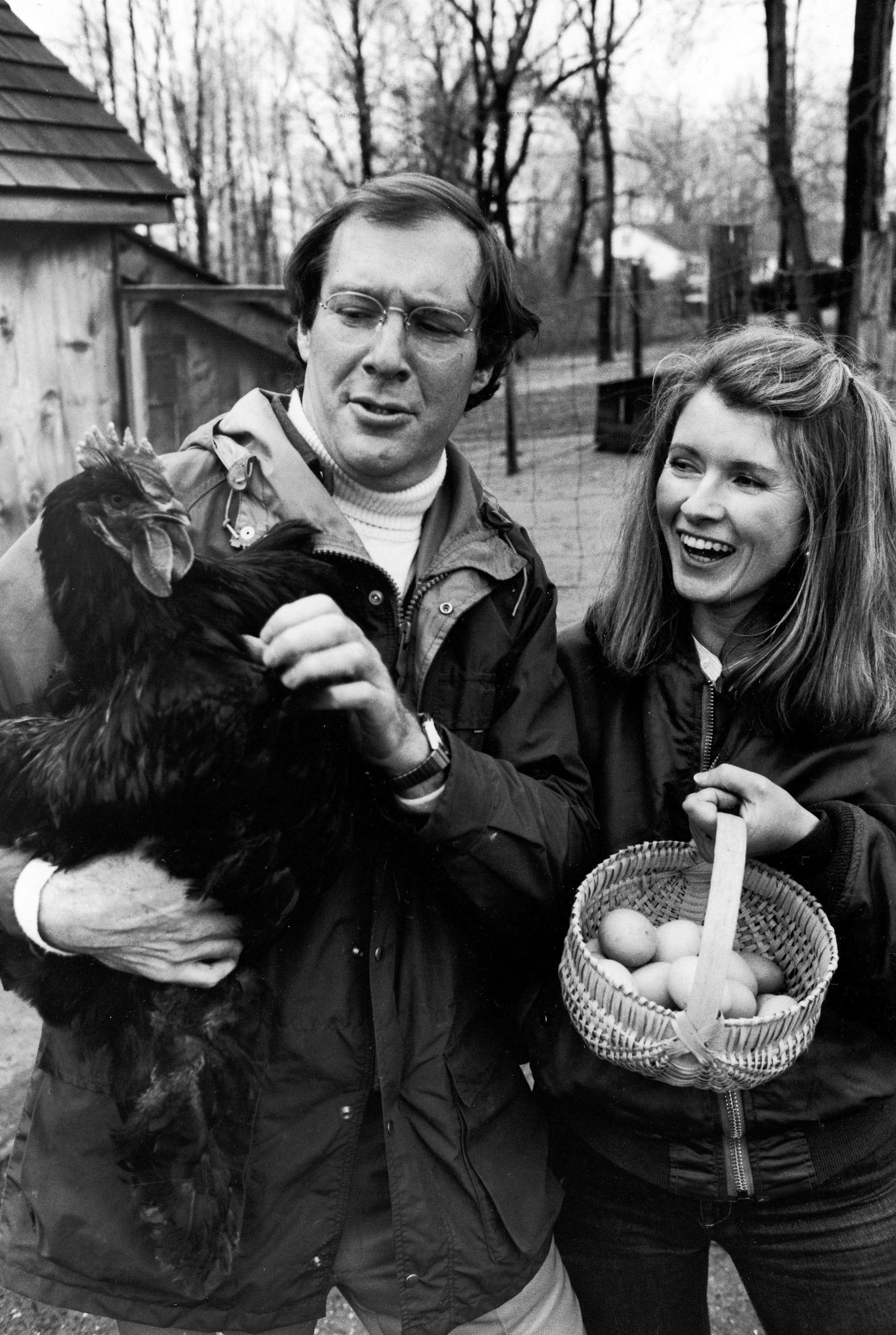 Andrew Stewart and Martha Stewart candidly pose at an outdoor farm. Andrew holds a rooster and Martha holds a basket of eggs