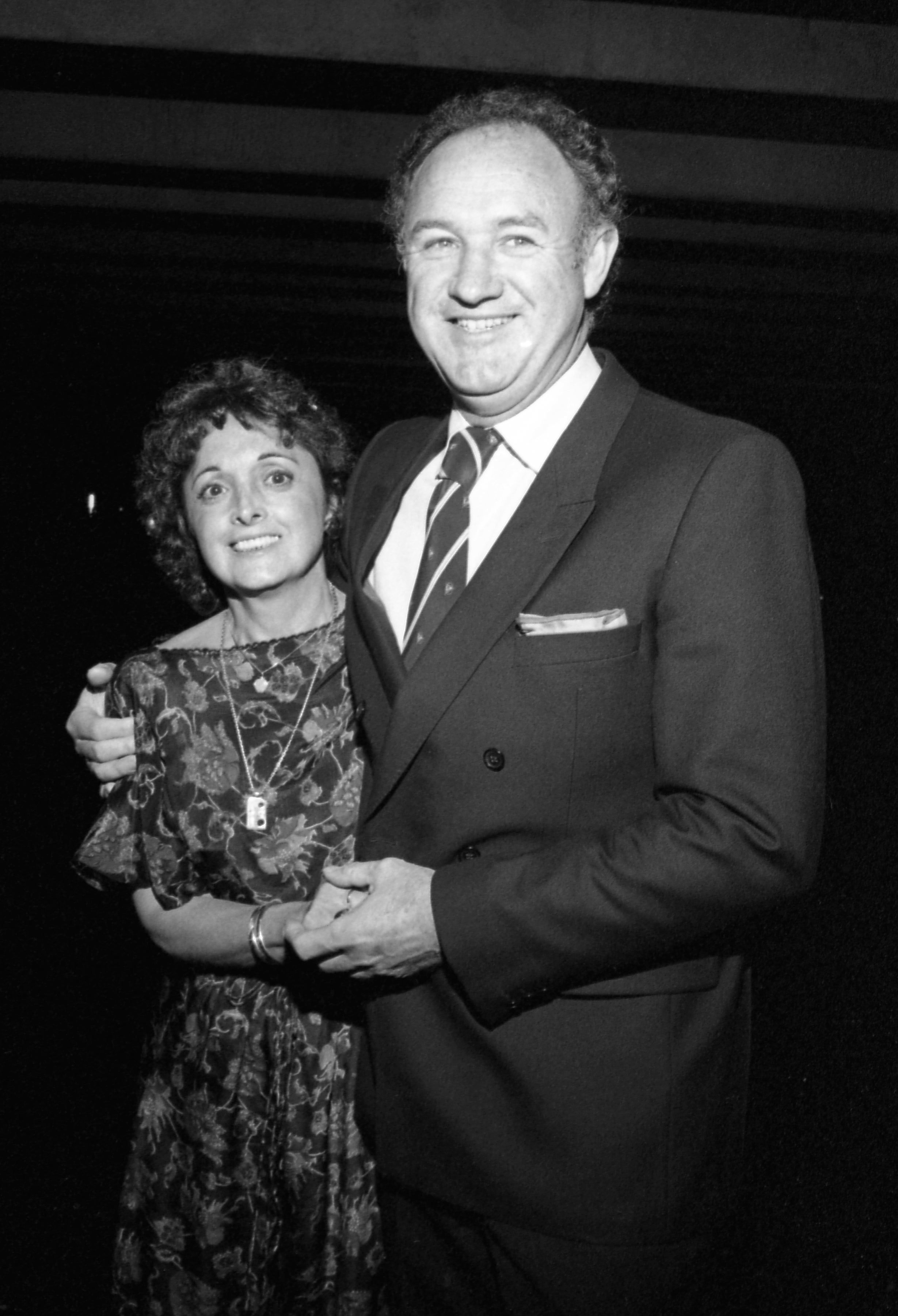 Faye Maltese and Gene Hackman pose together at an event with their arms around each other. Maltese wears a floral dress, and Hackman wears a suit and tie