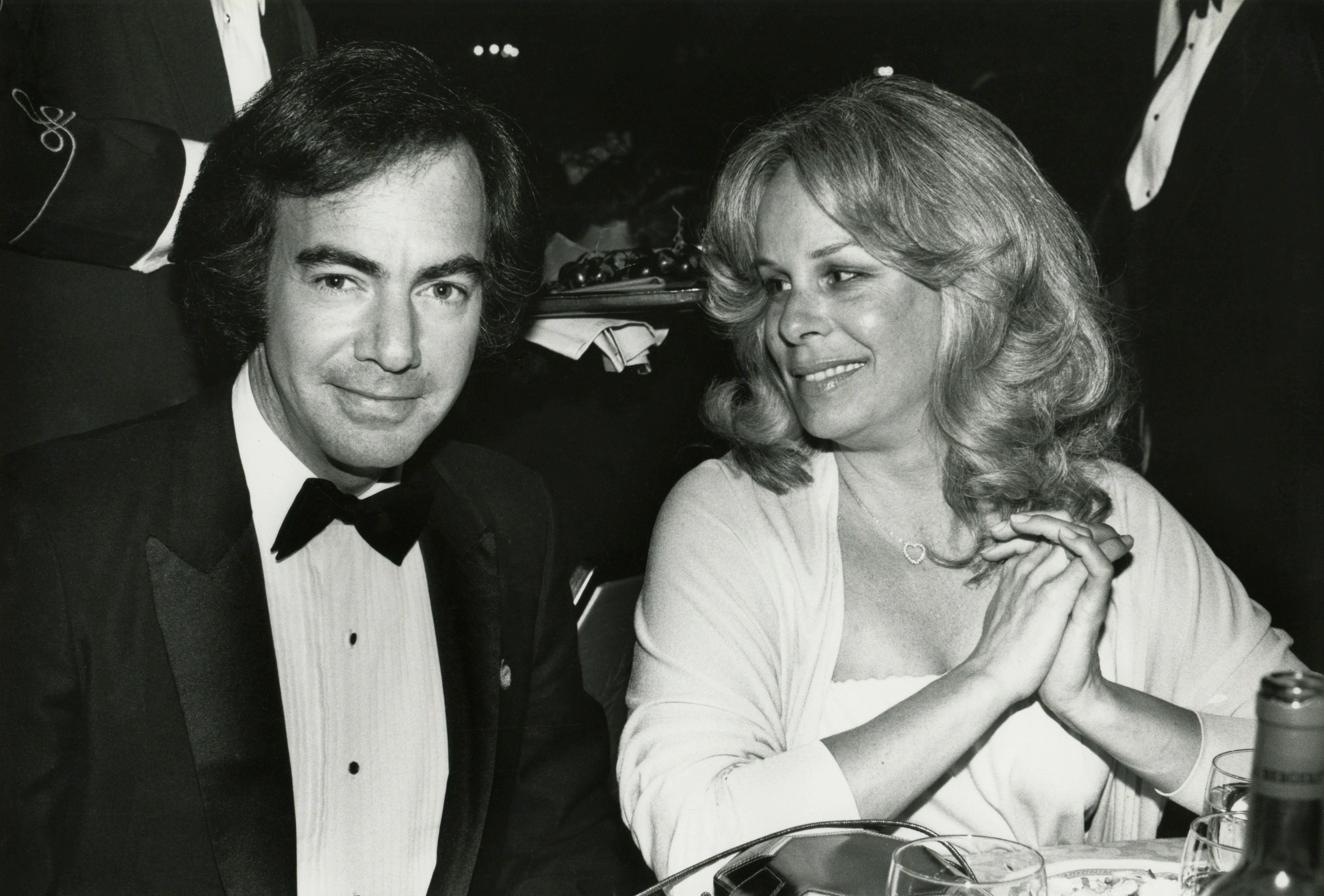 Neil Diamond and Marcia Murphey are seated at a formal event. Diamond wears a tuxedo, and Murphey wears a light dress. Both are smiling and engaged in conversation