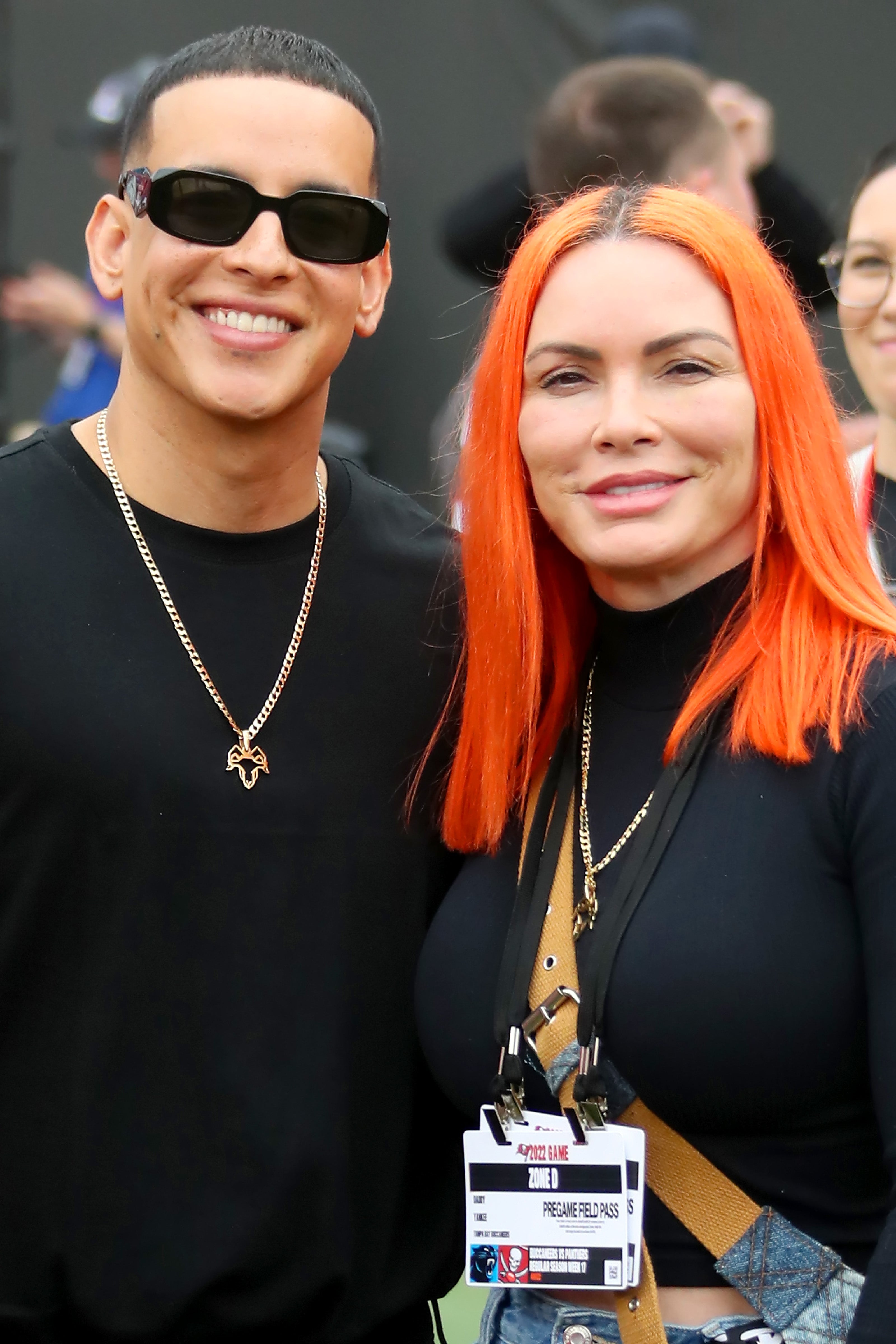 Daddy Yankee (Ramón Luis Ayala Rodríguez) and Mireddys González pose at an event. Daddy Yankee wears a black T-shirt, a gold necklace, and black sunglasses. González wears a black shirt and has bright orange hair