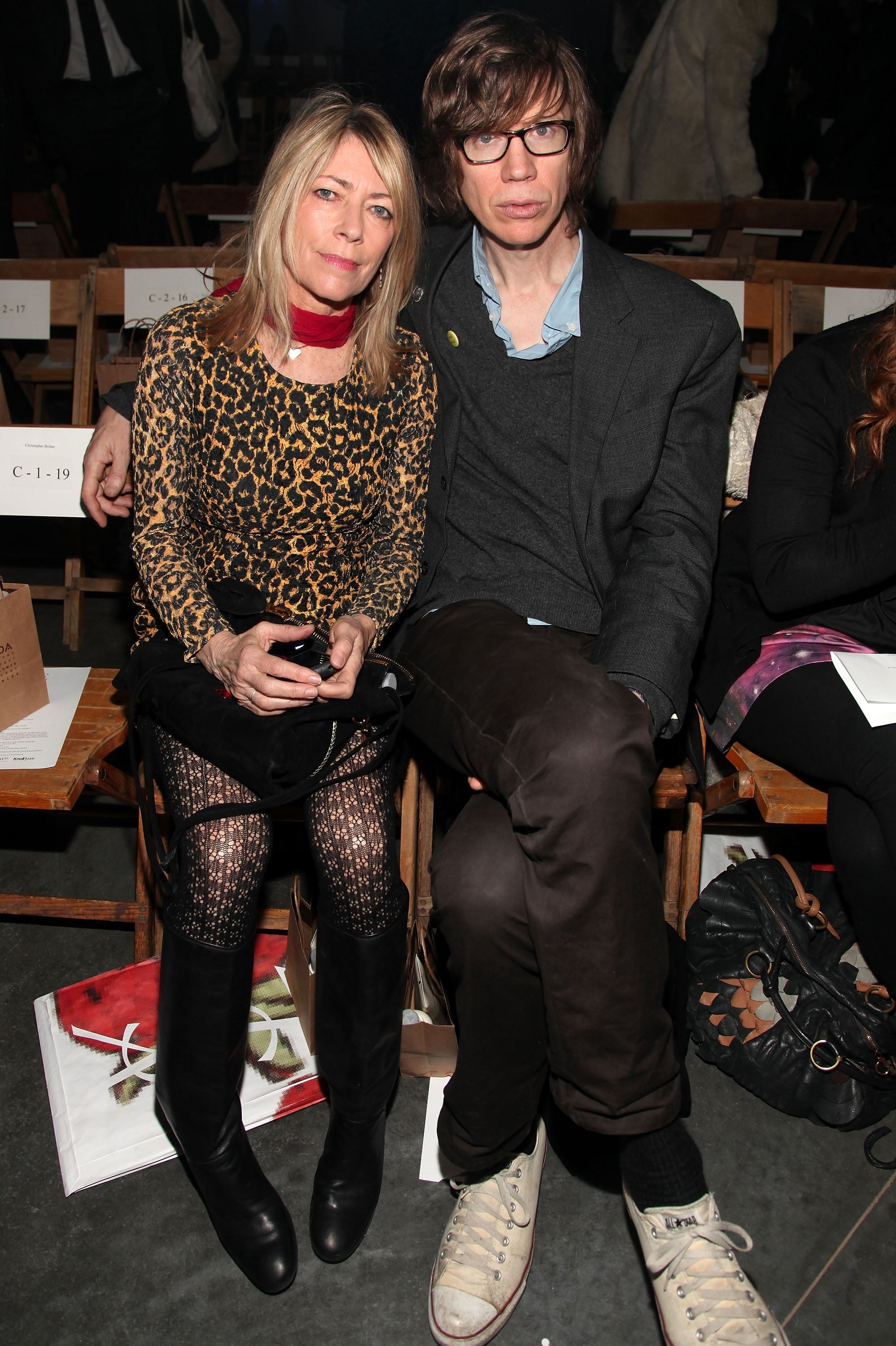 Kim Gordon and Thurston Moore sit side by side at an event. Gordon wears a cheetah-print shirt and patterned tights. Moore wears white Converse shoes, black pants, and glasses