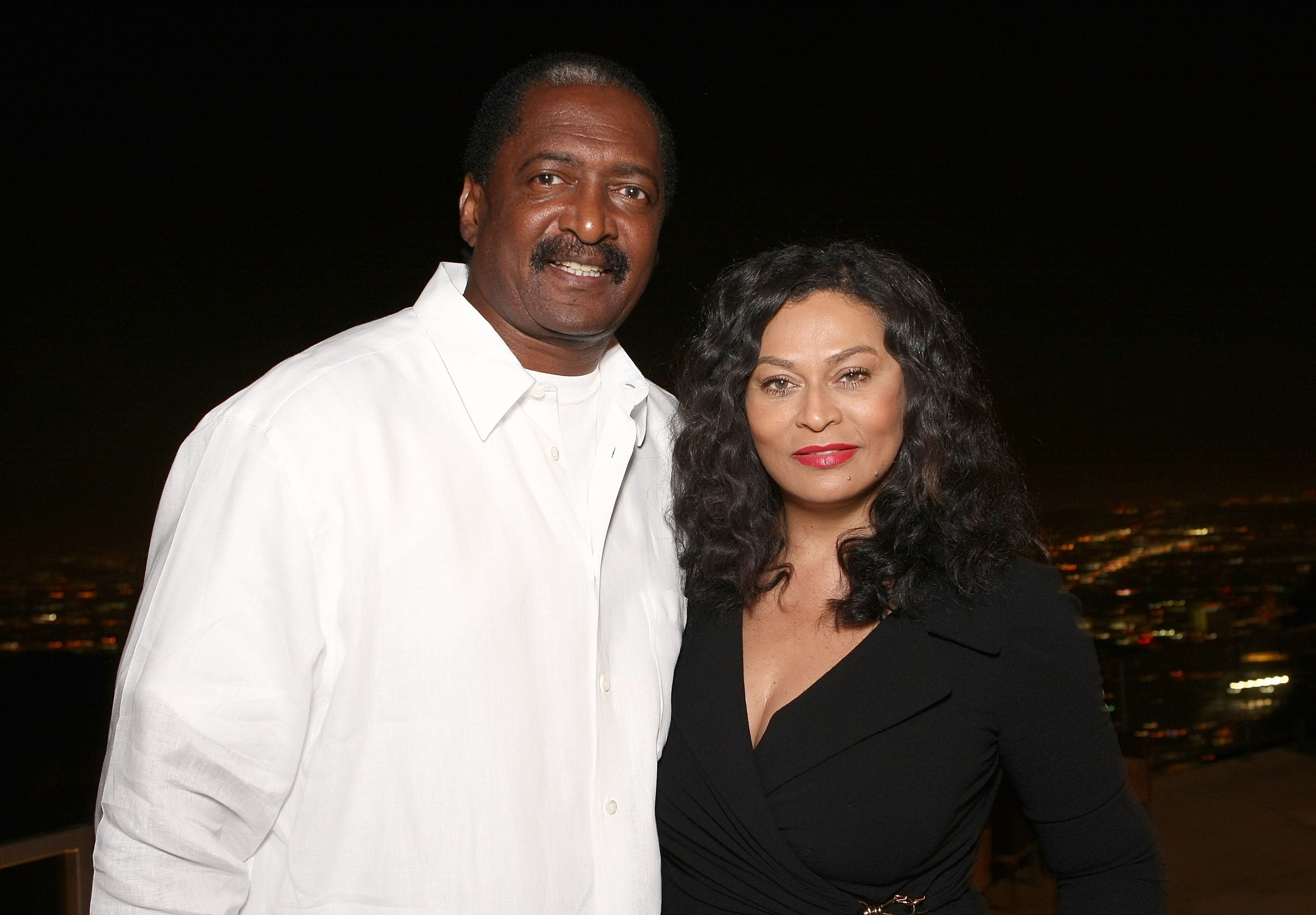 Matthew Knowles and Tina Knowles pose together at an event. Matthew wears a white button-up shirt, and Tina wears a black dress
