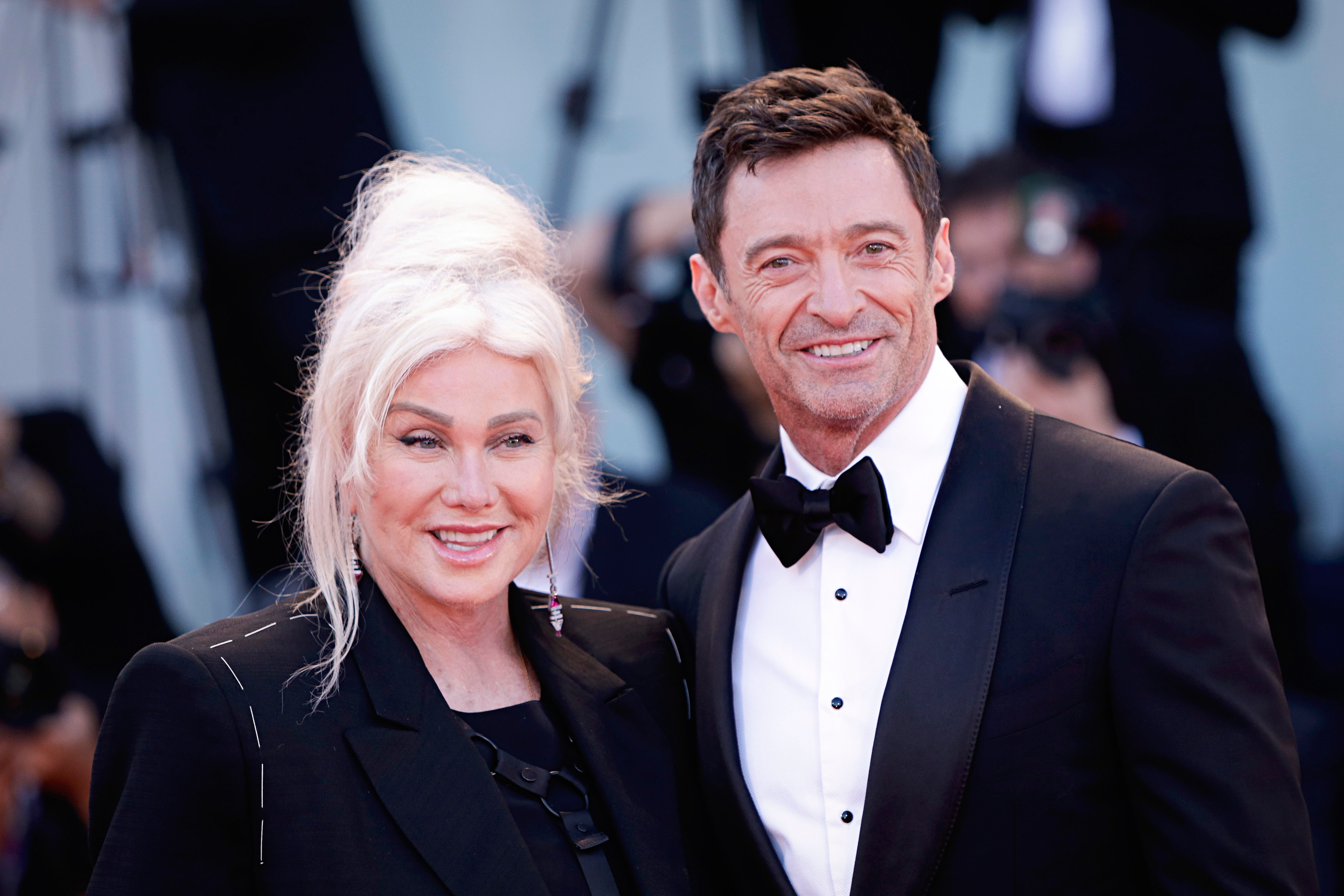 Deborra-lee Furness and Hugh Jackman pose on a red carpet. Furness wears an elegant updo with a black suit, and Jackman wears a classic tuxedo and bow tie