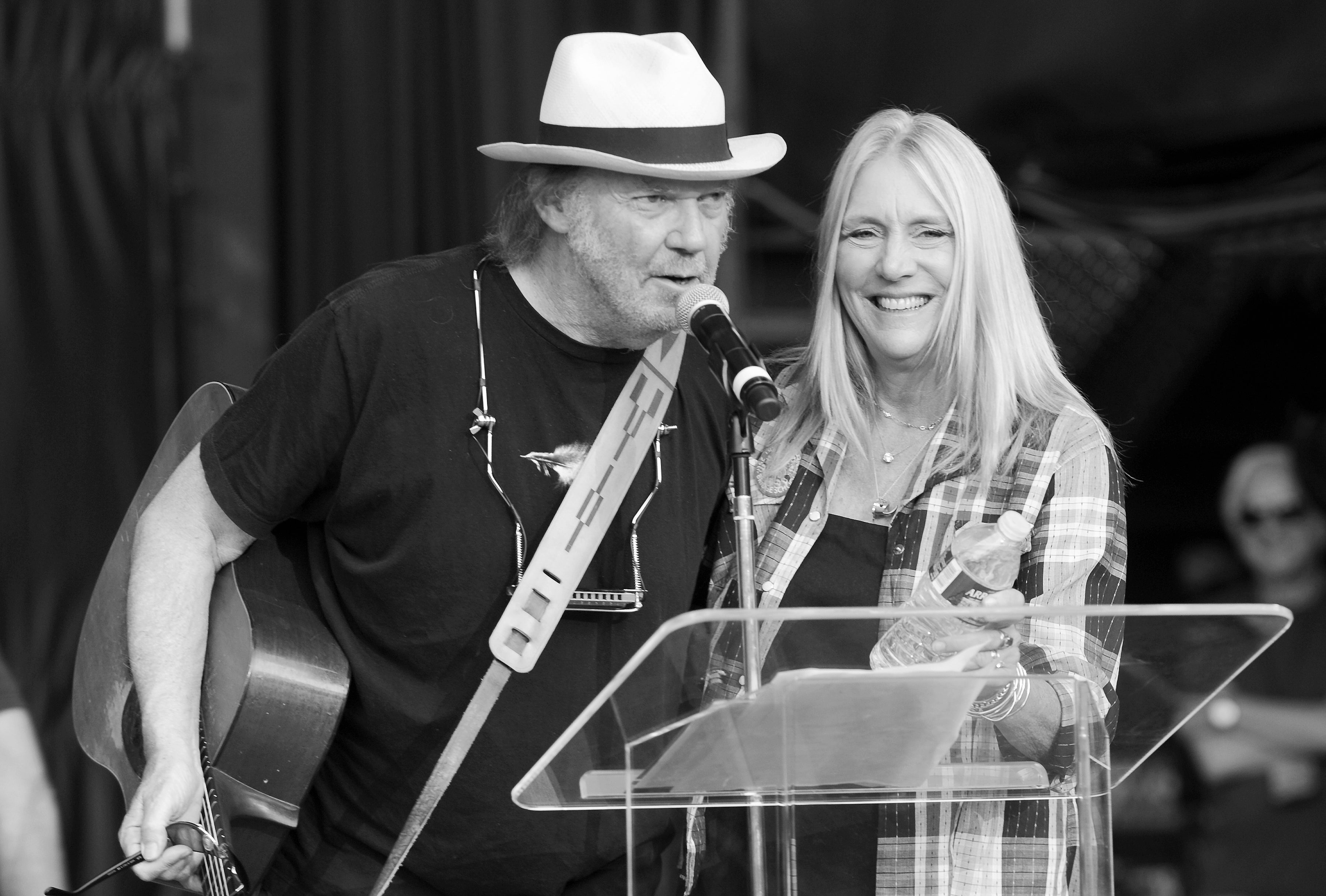 Neil Young and Pegi Young perform together on stage. Neil wears a black T-shirt and hat, and Pegi wears a plaid flannel. Neil speaks into a microphone while Pegi holds a bottle of water