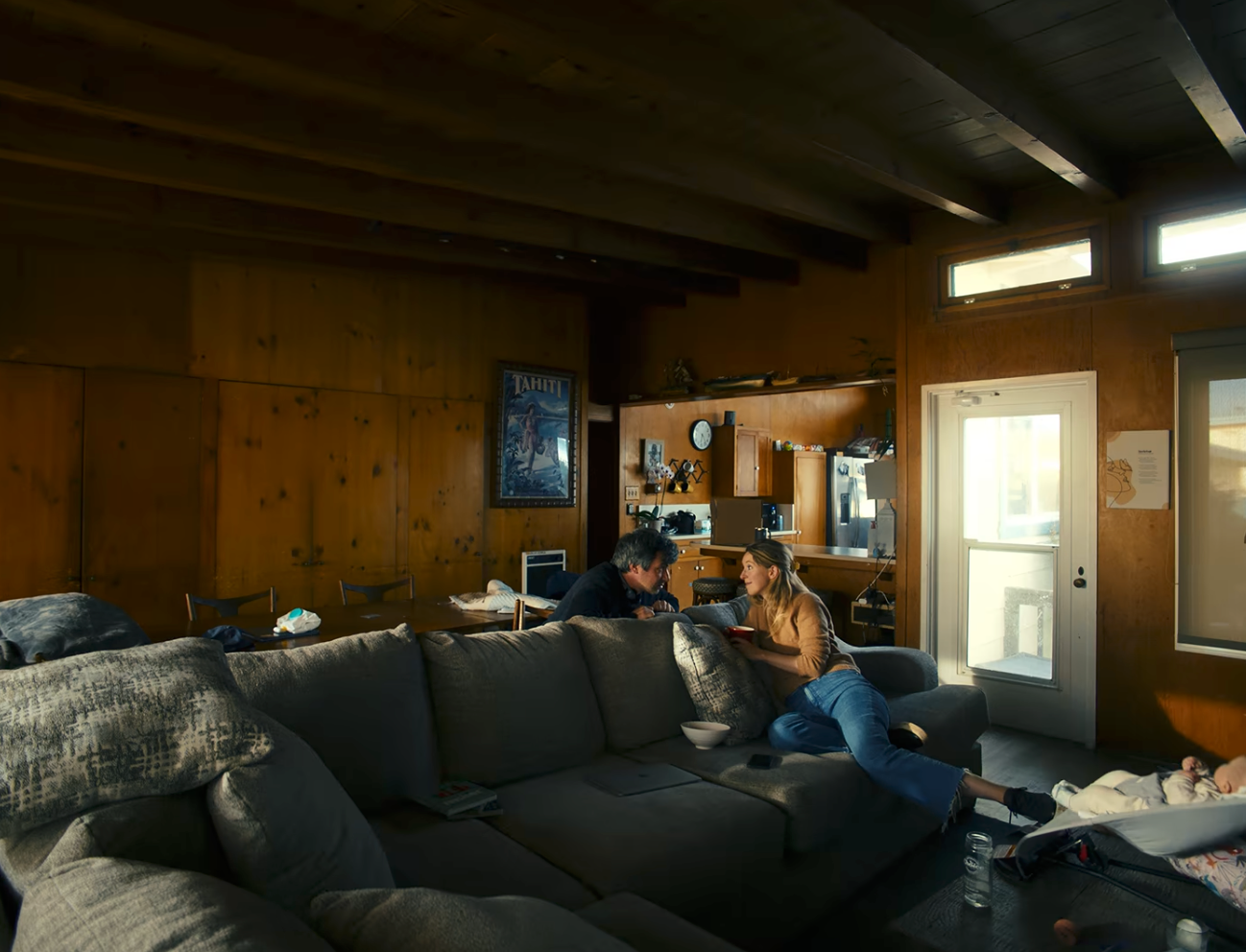 To personer sitter ved et bord i et koselig rom med trepanel og er i samtale. En sofa og en sovende baby i en dørvakt er i forgrunnen