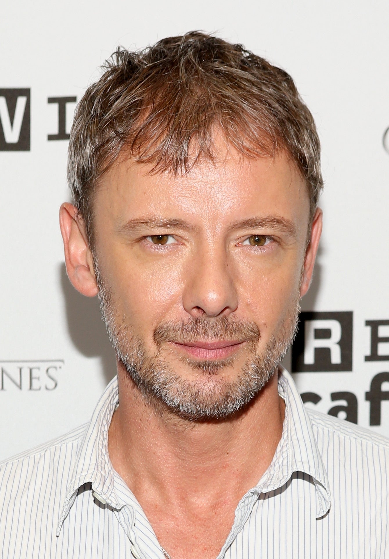 A person with short hair and a beard, wearing a striped shirt, attends an event, standing in front of an event backdrop