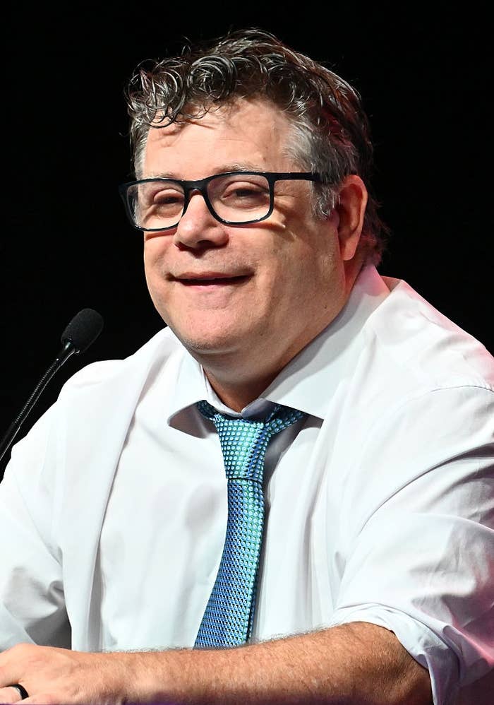 A person with glasses and a blue tie is speaking into a microphone, smiling
